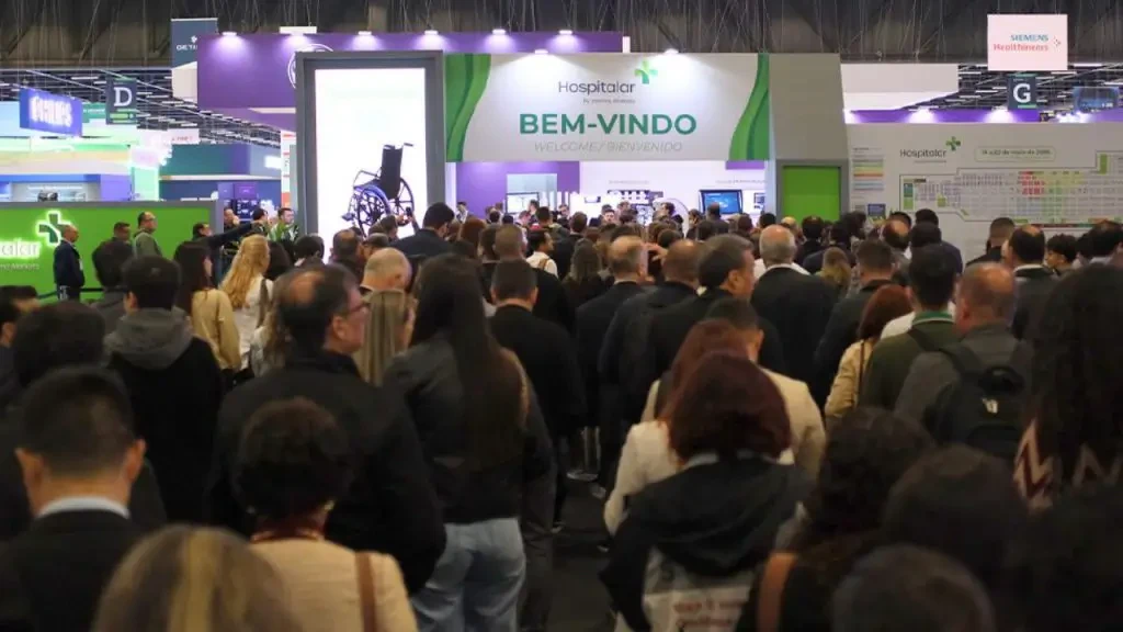 Público de profissionais caminhando pela entrada da feira Hospitalar em pavilhão de exposições de saúde.