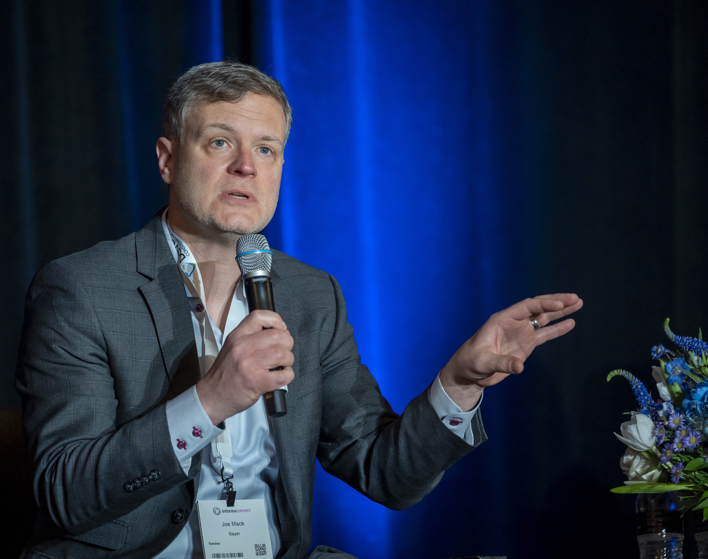 A speaker speaks into a microphone, behind him is a blue curtain.