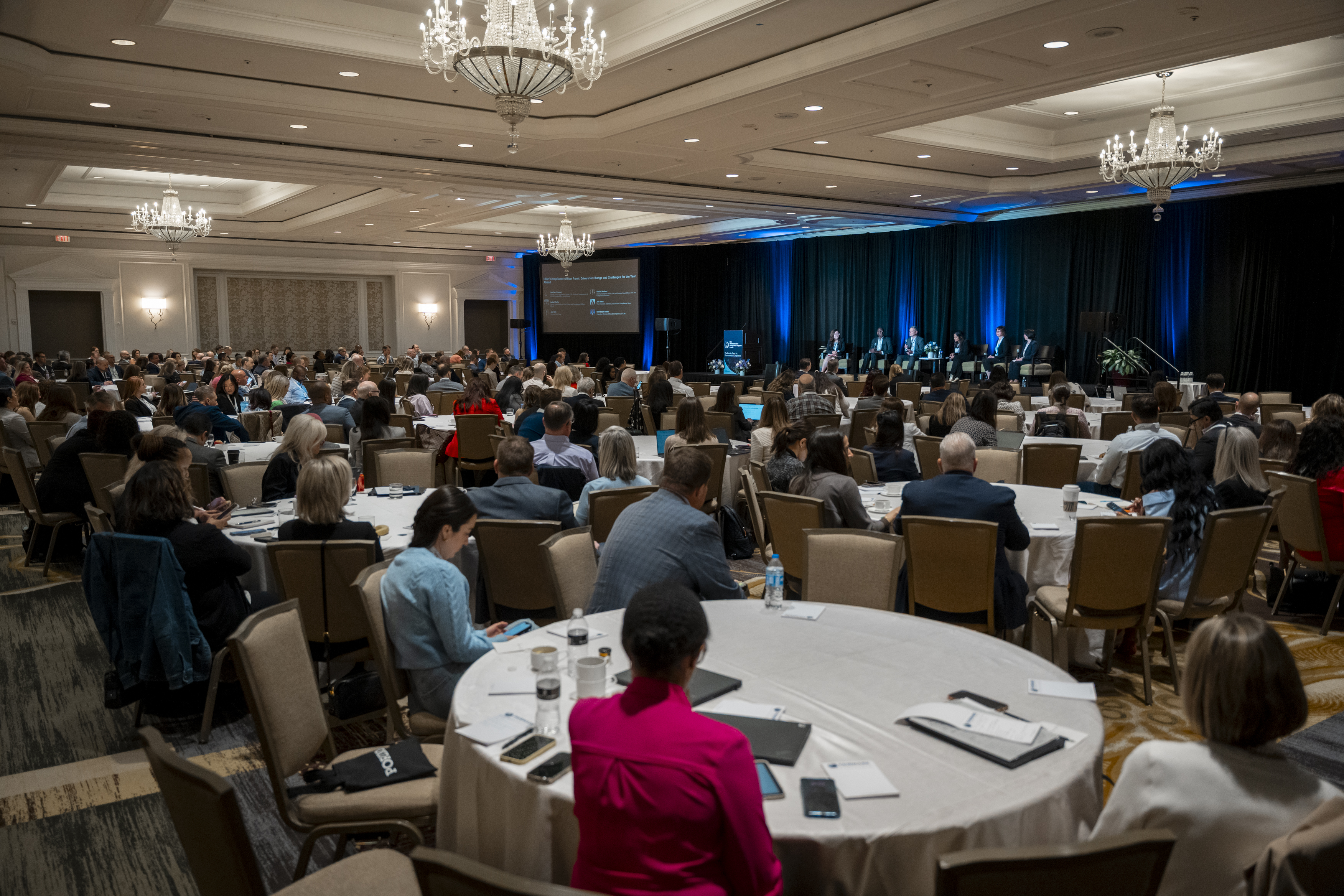 A busy conference room with many round tables and attendees sitting whilst a panel sits at the front.
