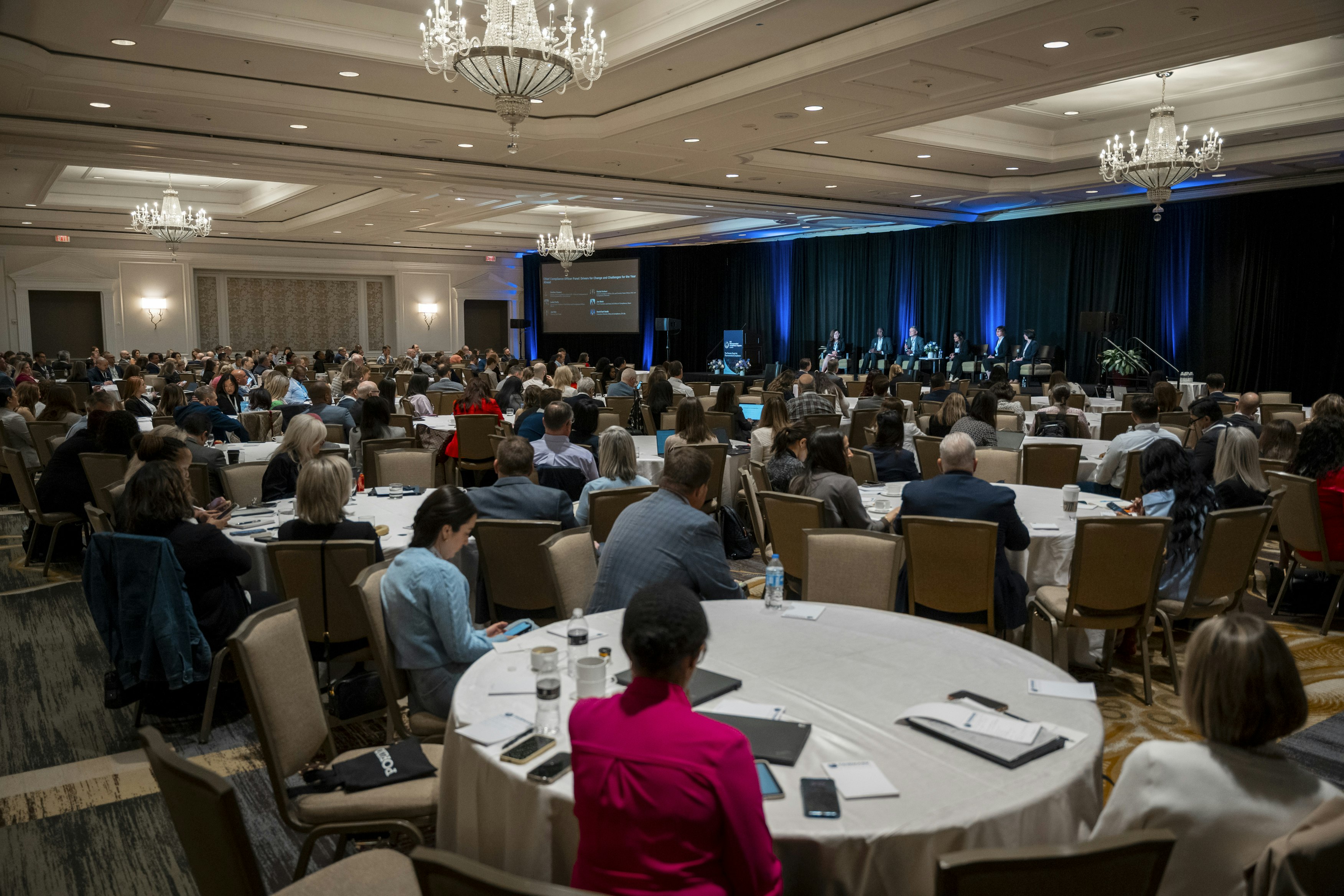 A busy conference room with many round tables and attendees sitting whilst a panel sits at the front.