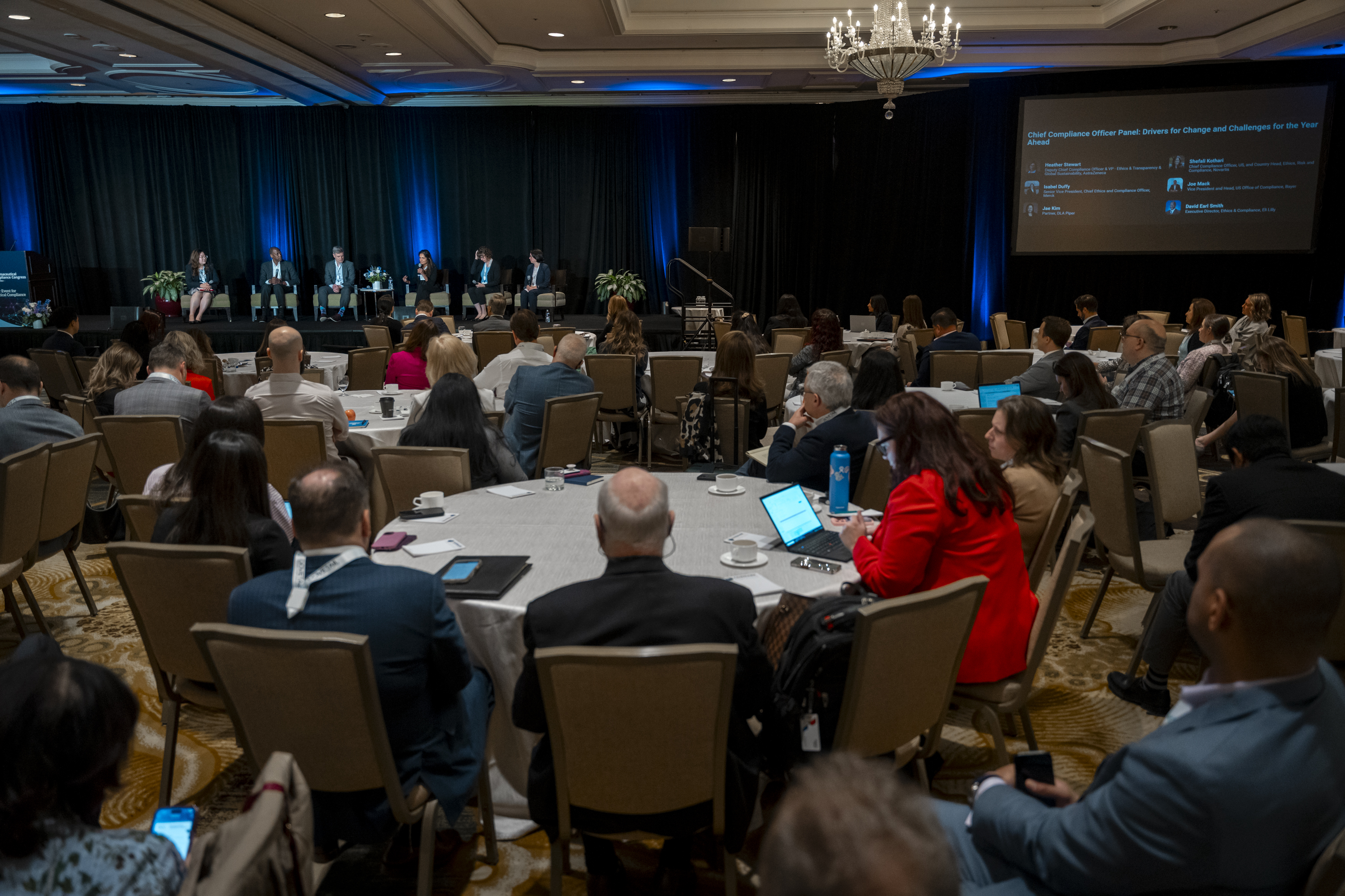 Conference room full of attendees sat on round tables whilst a panel of six speakers talk on stage.