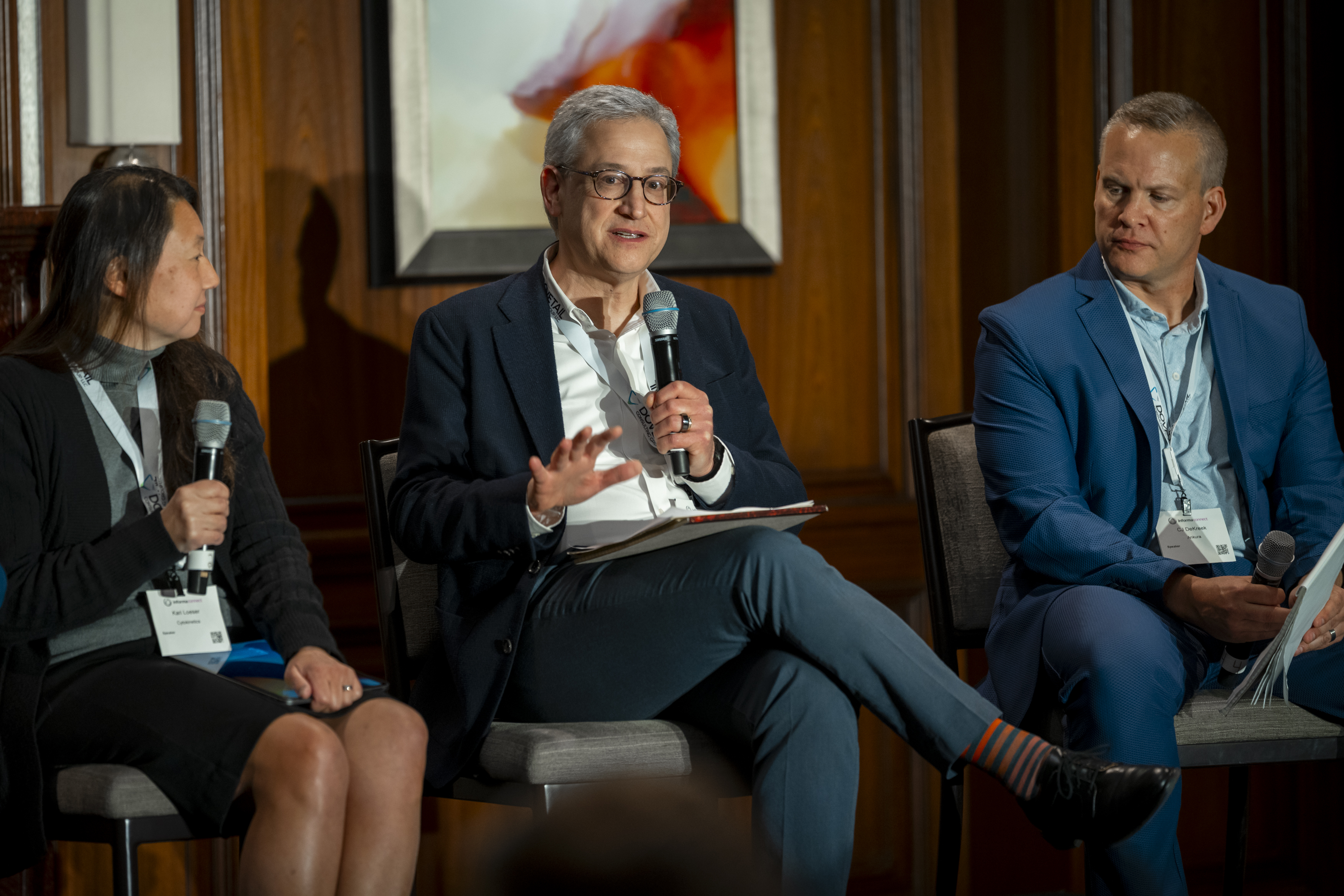 Three speakers sit on stage with microphones in hands, one sat in the middle is speaking.