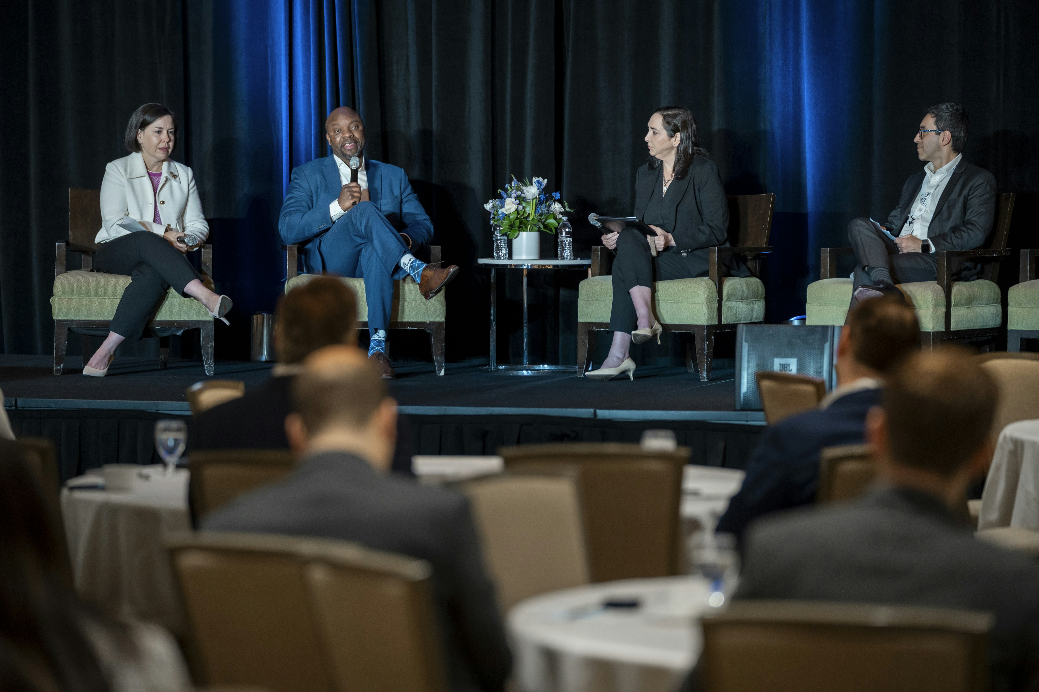 Four speakers sit on a panel on stage in the midst of conversation. In the background attendees sit on round tables.