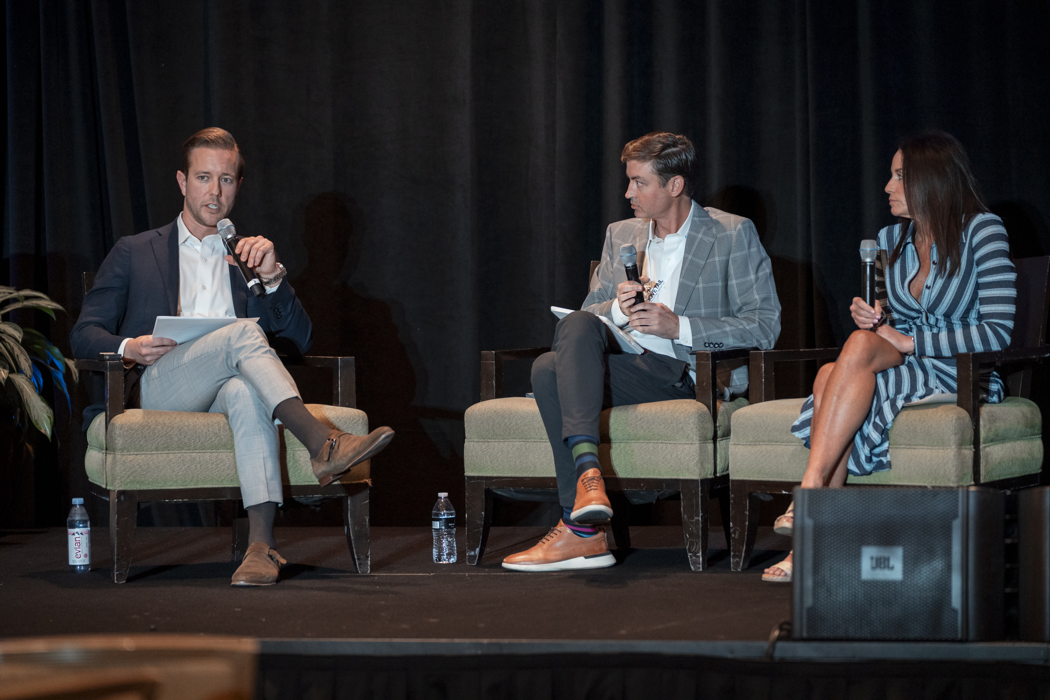 Three speakers sit on stage with microphones in hand, one is talking.
