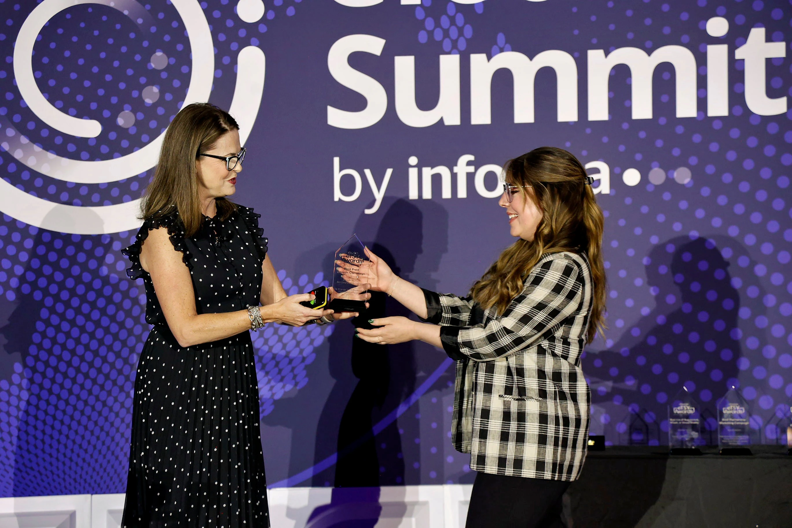 Two women accepting an eTSY Award. The woman on the left has dark curly hair and is wearing a black shirt with grey jeans. The woman on the right has straight blonde hair and is wearing a white blouse, black pants, and an Apple Watch.