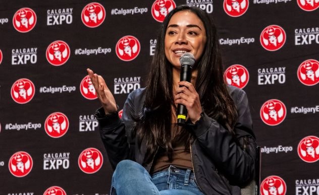 Aimee Garcia smiles as she speaks into a microphone while wearing a black jacket