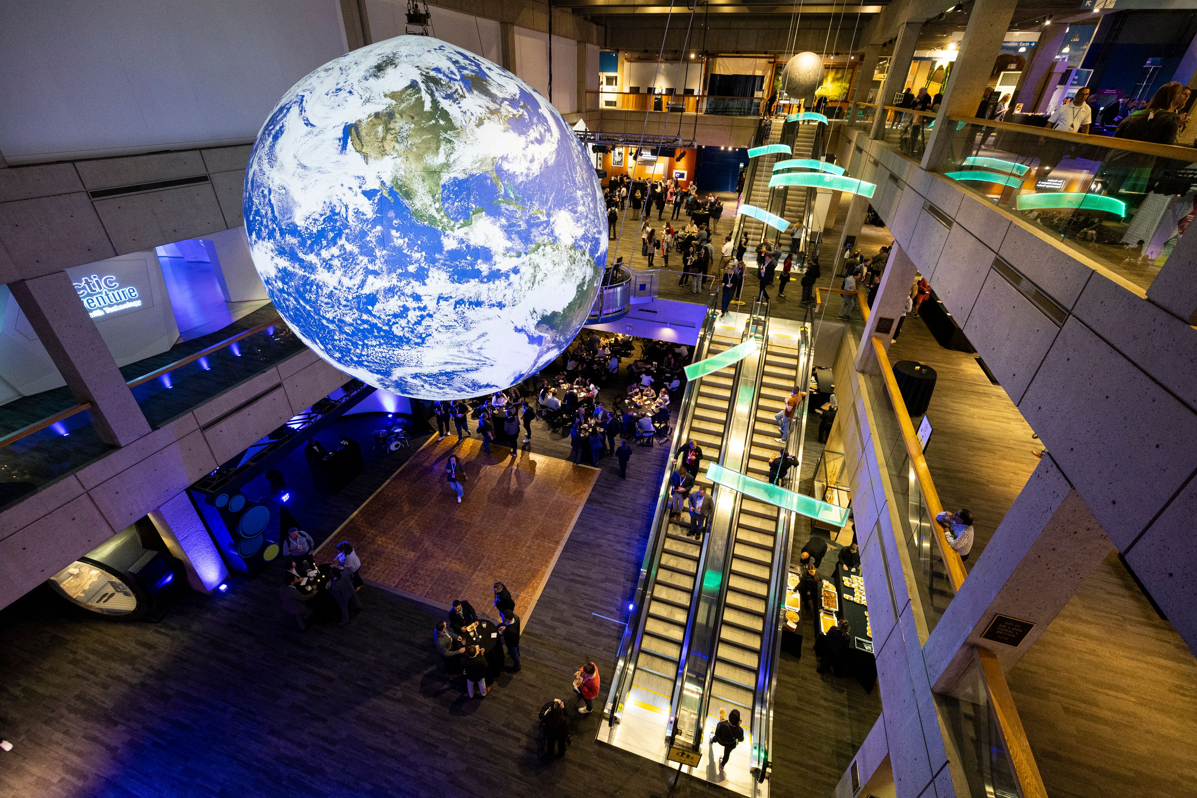 A large illuminated world globe hangs from the ceiling in the exhibition centre with people on escalators on the ground and first floors.
