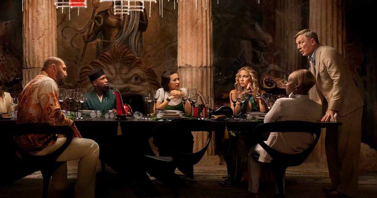 A screen cap from Glass Onion: A Knives Out Mystery featuring several characters all seated/standing by a table. From left to right: Duke Cody (Dave Bautista), Lionel Toussaint (Leslie Odom Jr.), Peg (Jessica Henwick), Birdie Jay (Kate Hudson), and Benoit Blanc (Daniel Craig).