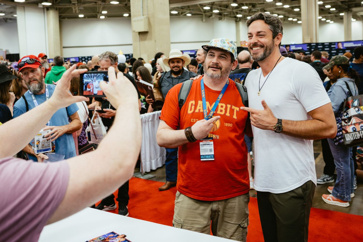 A fan in a red Gambit t-shirt and colorful cap poses with Zachary Levi, who wears a white t-shirt, while someone takes their photo on a phone. Both are smiling and pointing at each other as a crowd fills the convention floor behind them.