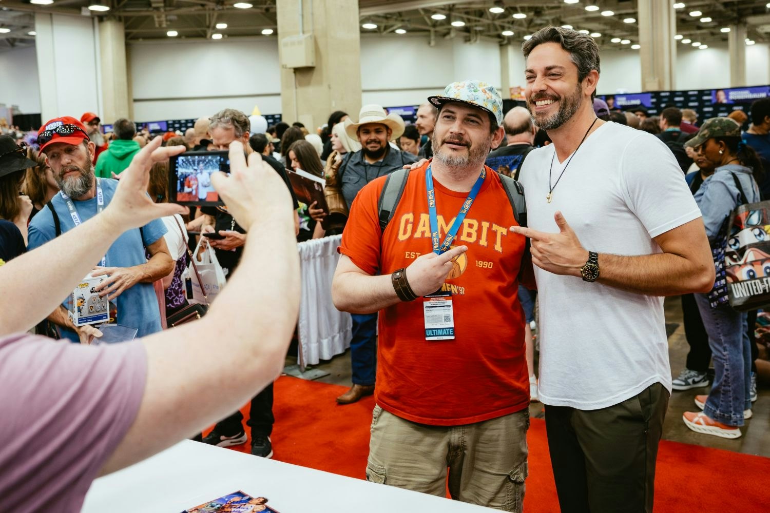 A fan in a red Gambit t-shirt and colorful cap poses with Zachary Levi, who wears a white t-shirt, while someone takes their photo on a phone. Both are smiling and pointing at each other as a crowd fills the convention floor behind them.