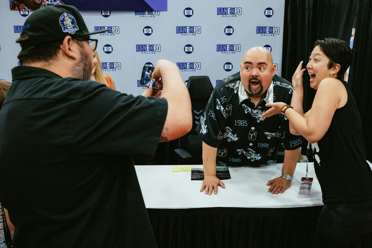 Comedian Gabriel “Fluffy” Iglesias leans dramatically on a signing table, wide-eyed with his mouth open, matching the playful expression of a fan beside him. Another fan captures the candid moment with their phone.