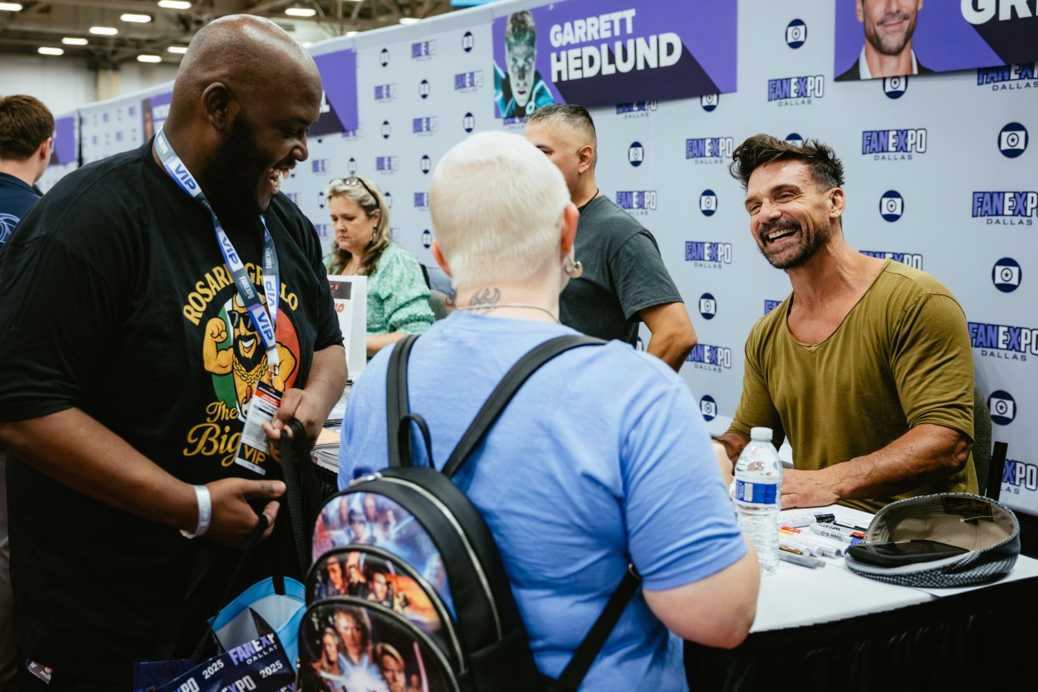 Actor Frank Grillo, seated at his signing table, shares a laugh with two fans—one in a “Rosario’s Big Dog” t-shirt and the other with a platinum buzz cut and Star Wars-themed backpack. The banner above reads “Garrett Hedlund,” indicating a shared signing area.