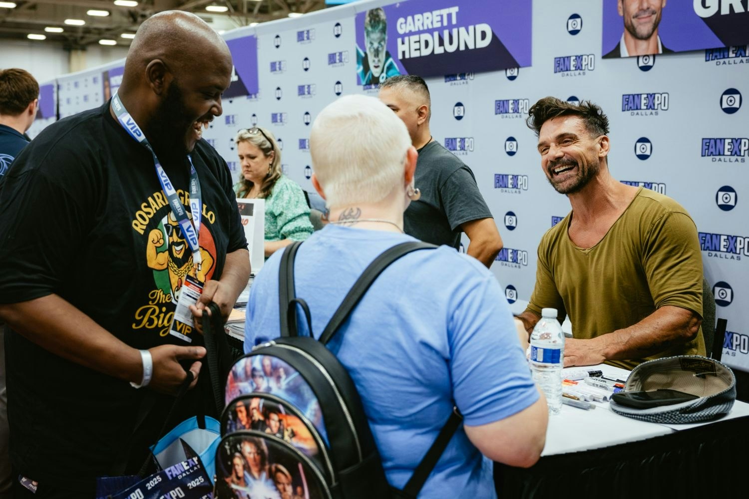Actor Frank Grillo, seated at his signing table, shares a laugh with two fans—one in a “Rosario’s Big Dog” t-shirt and the other with a platinum buzz cut and Star Wars-themed backpack. The banner above reads “Garrett Hedlund,” indicating a shared signing area.