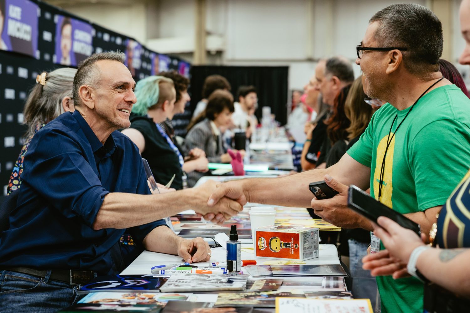 Actor Hank Azaria smiles warmly as he shakes hands with a fan wearing a green t-shirt. The table is covered in colorful autograph prints and markers, and several other celebrities can be seen seated further down the signing row.