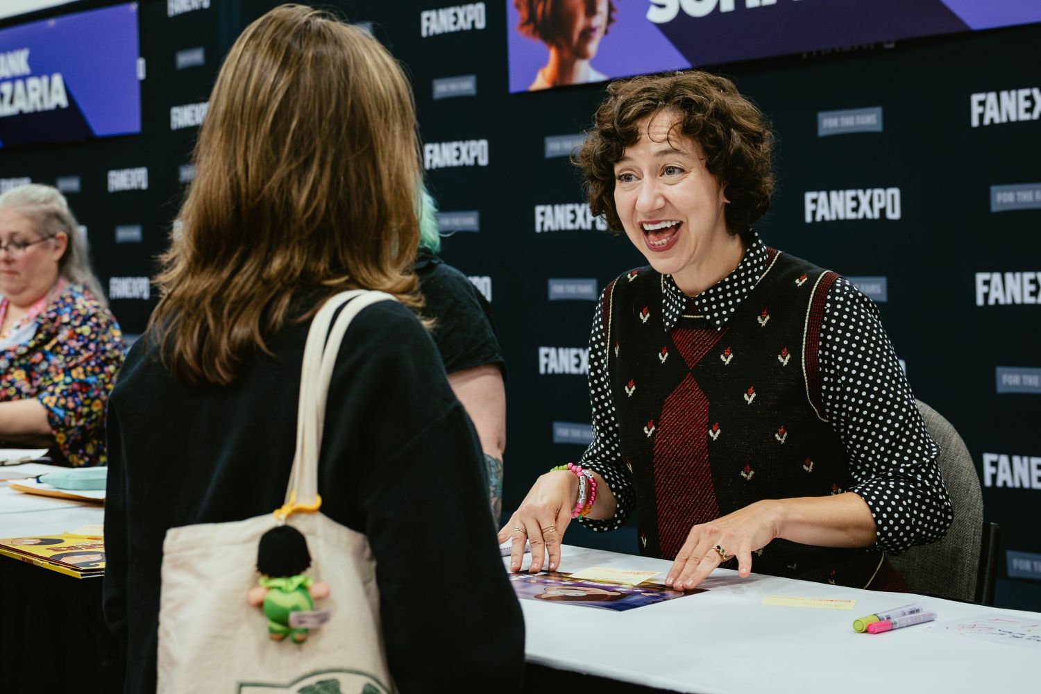 Actress Kristen Schaal, seated at her autograph table, beams with excitement while talking to a fan. She wears a patterned sweater vest and rests her hands on a glossy autograph print. Her FAN EXPO Dallas nameplate is visible behind her.
