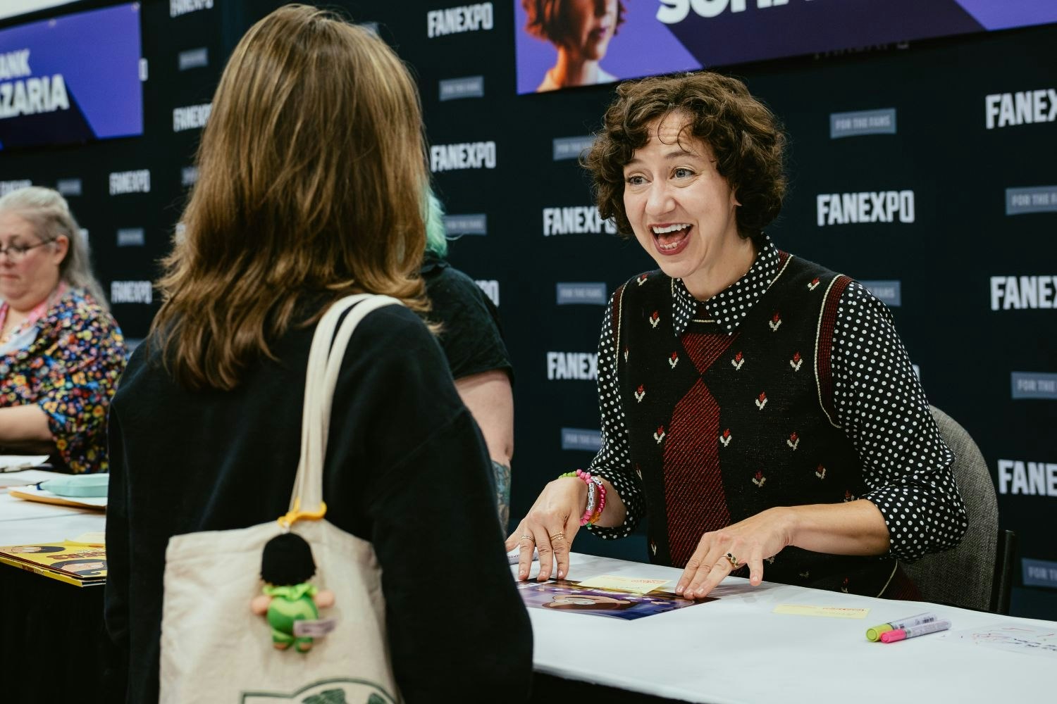 Actress Kristen Schaal, seated at her autograph table, beams with excitement while talking to a fan. She wears a patterned sweater vest and rests her hands on a glossy autograph print. Her FAN EXPO Dallas nameplate is visible behind her.