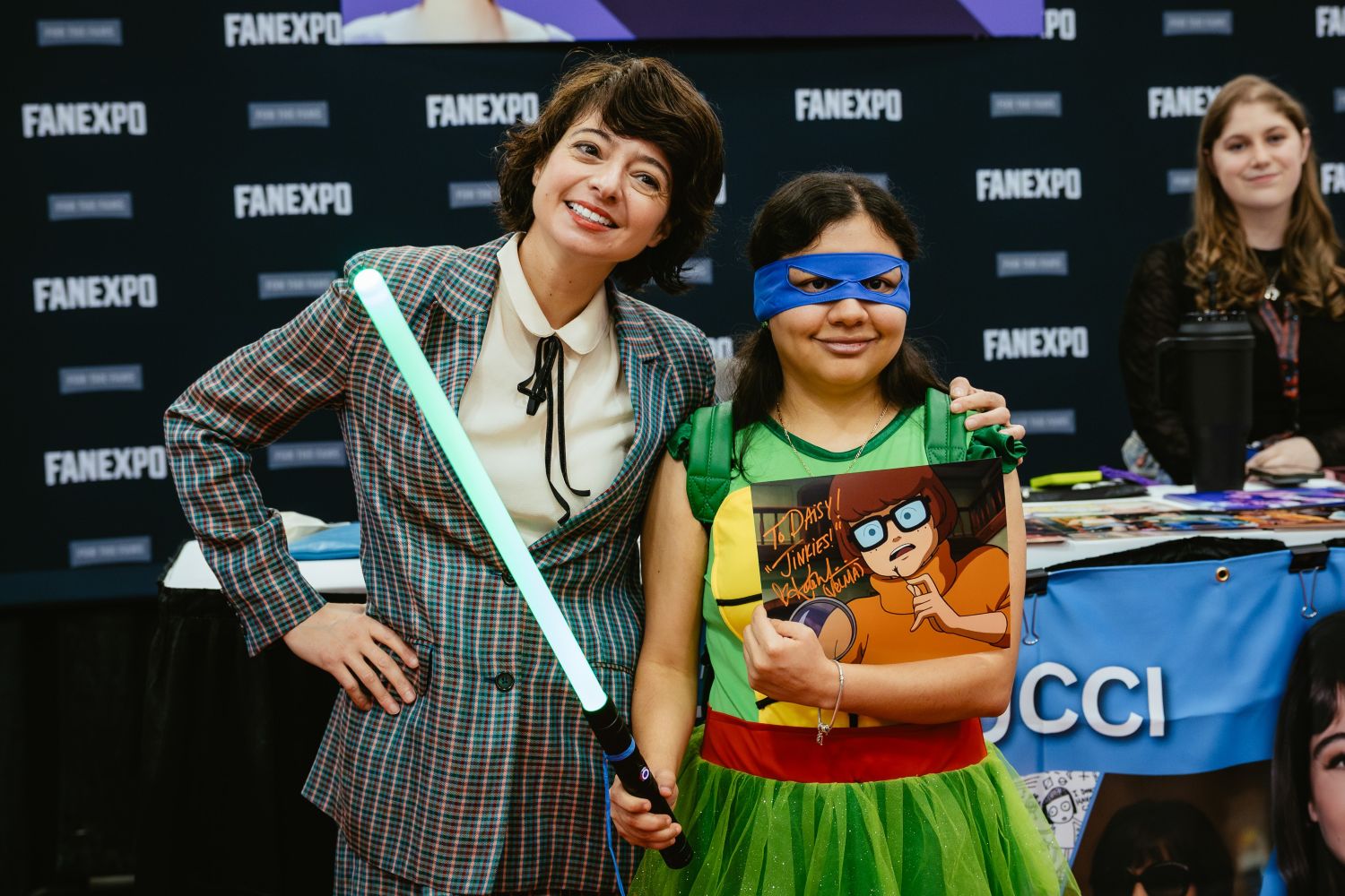 Actress Kate Micucci poses with a young fan dressed as a Teenage Mutant Ninja Turtle and holding a Velma Dinkley autograph, while holding a glowing lightsaber. Both smile brightly for the camera in front of her FAN EXPO banner.