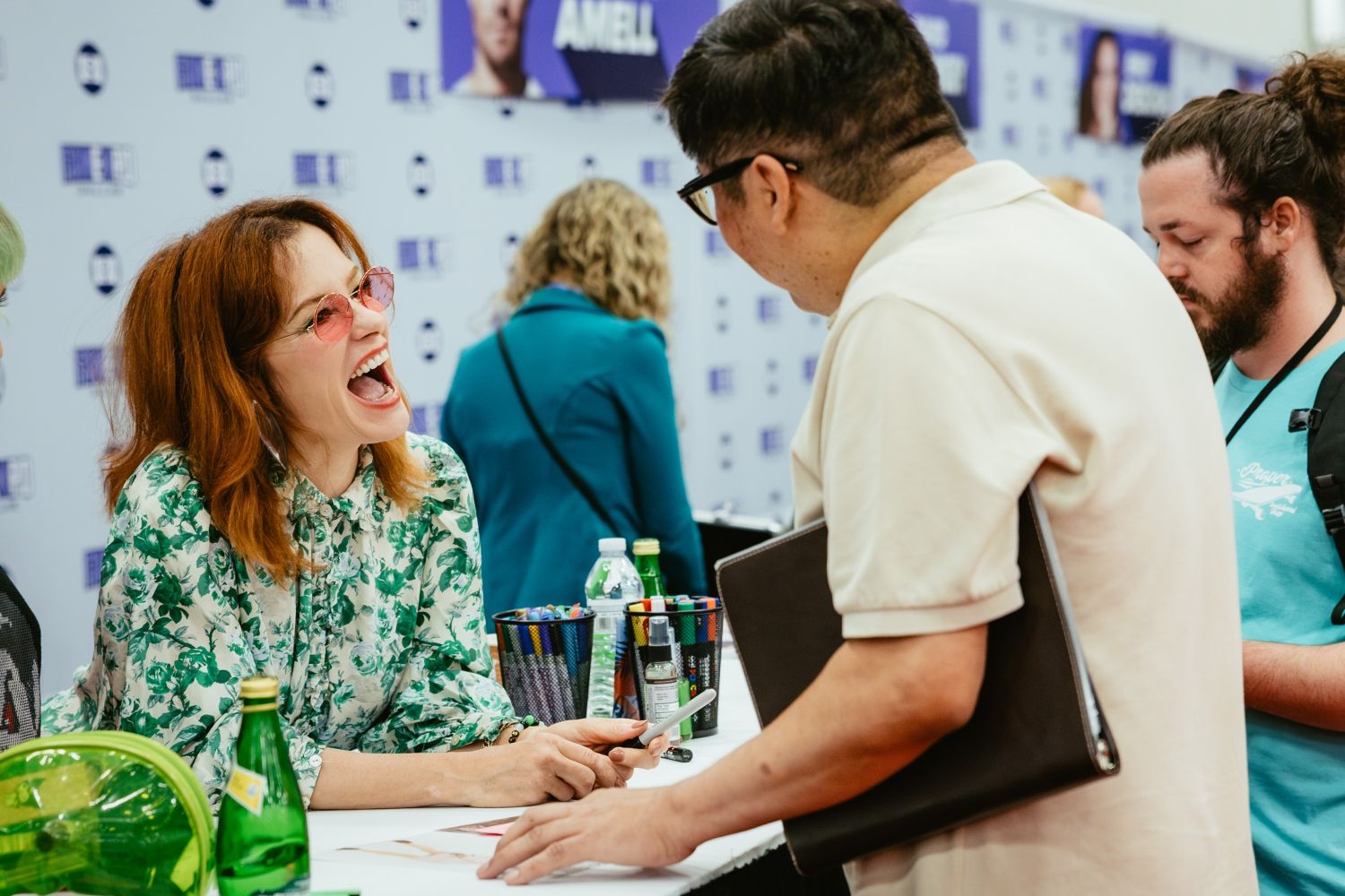 Actress Parker Posey laughs with a fan across her signing table. She wears a floral blouse and pink sunglasses, holding a pen and gesturing enthusiastically while chatting. Colorful markers and a water bottle sit on the table.