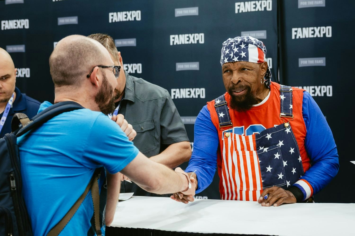 Mr. T, dressed in his signature red, white, and blue overalls and a matching bandana, shakes hands with a fan across his signing table. The fan wears glasses and a blue t-shirt, smiling as they meet the pop culture icon.