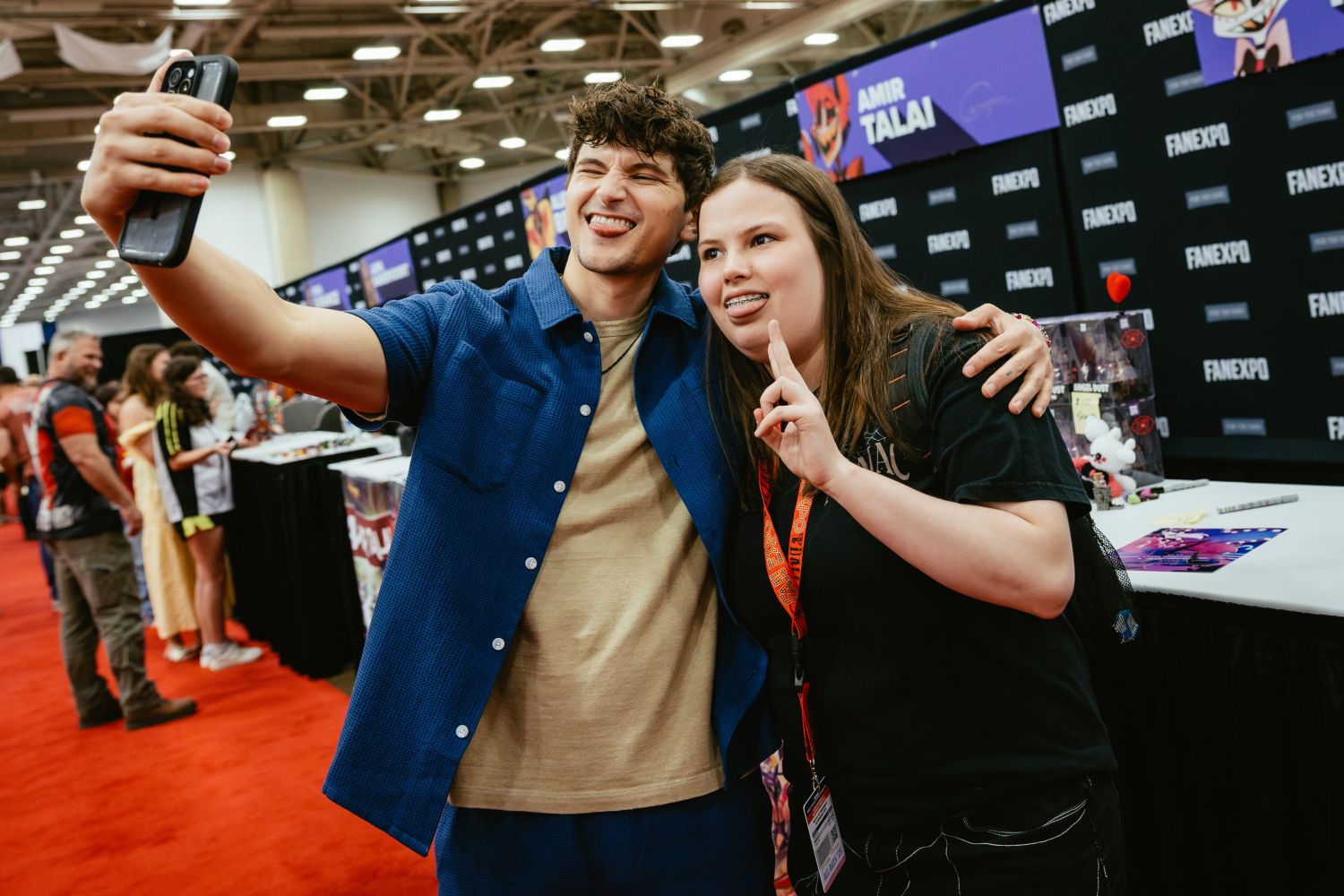 Voice actor Blake Roman, wearing a blue short-sleeve button-up and tan shirt, takes a cheerful selfie with a fan at a signing table. The fan, wearing a black shirt and a FAN EXPO lanyard, smiles and flashes a peace sign. The backdrop features branding for voice actor Amir Talai.