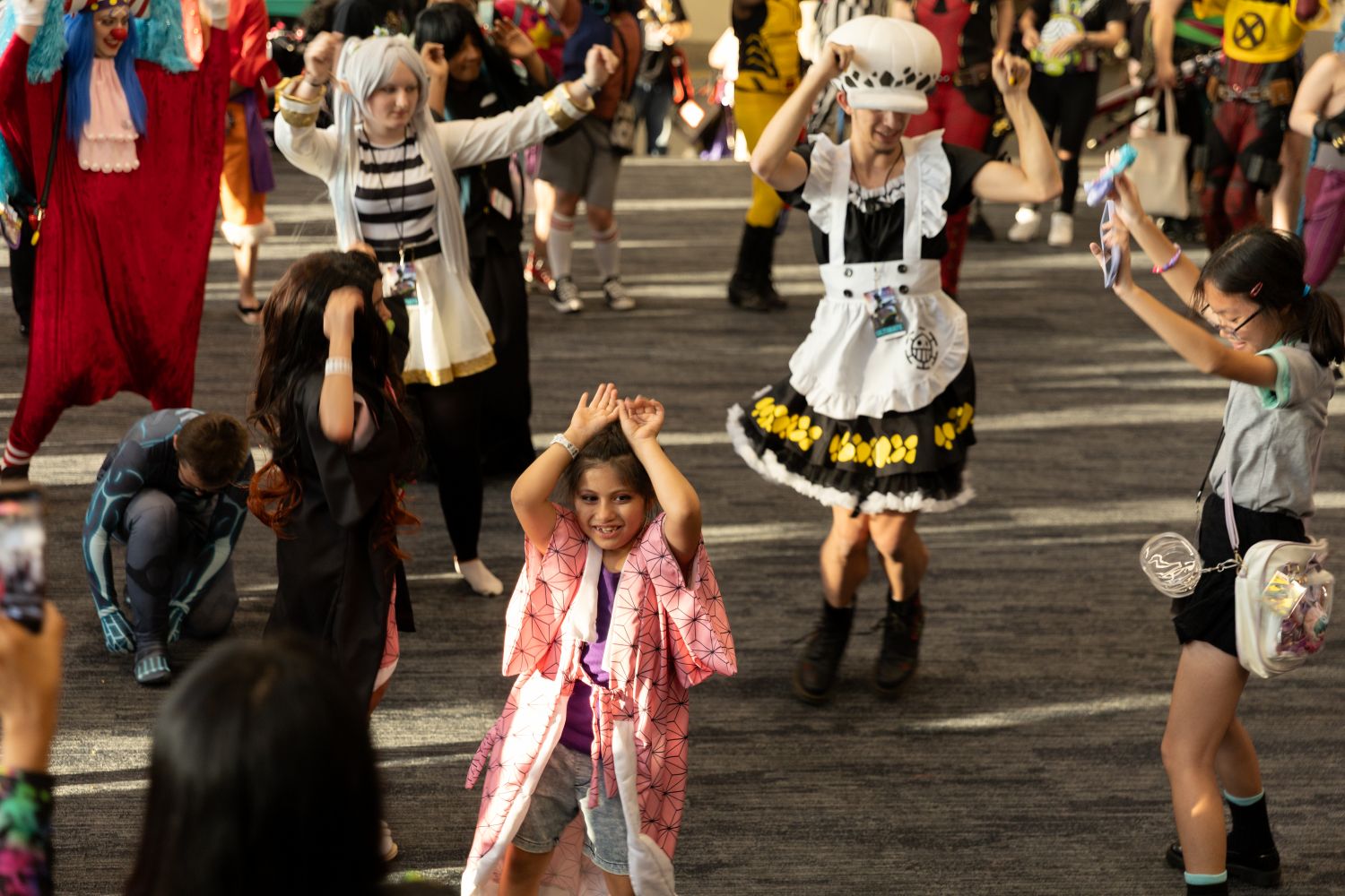 A young girl in a pink kimono-style cosplay leads a group dance during Anime After Dark. She smiles brightly with her arms raised, surrounded by fellow cosplayers in colorful and varied anime-inspired outfits.