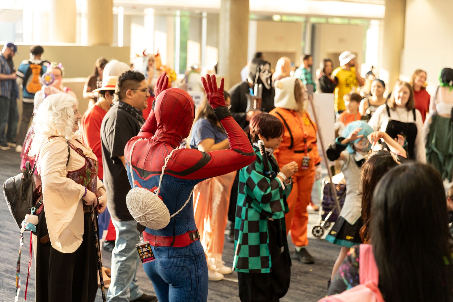 A crowd of cosplayers, including characters from Spider-Man, Demon Slayer, and Naruto, participate in a group dance under warm lighting. The atmosphere is lively and filled with joy as fans gather in costume.