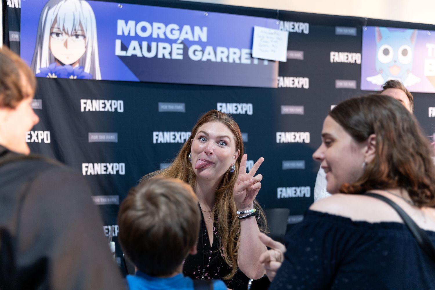 Voice actor Morgan Lauré Garrett makes a funny face while flashing a peace sign for a young fan in front of her autograph booth. The FAN EXPO Dallas backdrop and her guest banner are visible behind her.