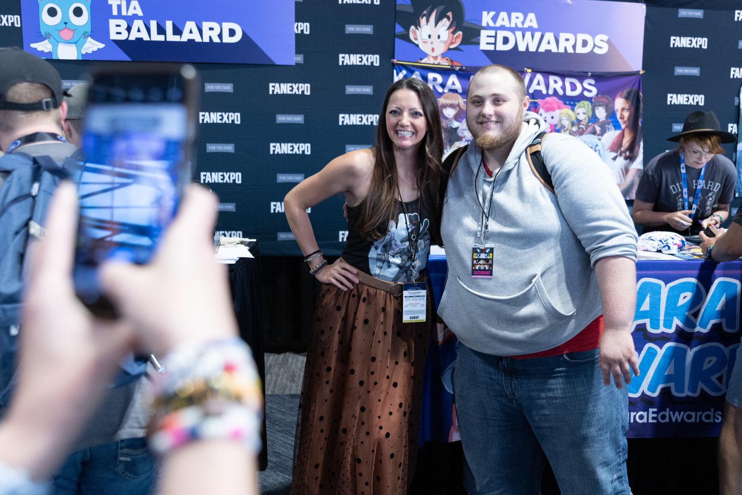 Voice actor Kara Edwards poses with a fan for a photo. She smiles brightly in a sleeveless black top and polka-dot skirt, while the fan in a gray hoodie beams beside her. Their photo is being snapped by someone in the foreground.