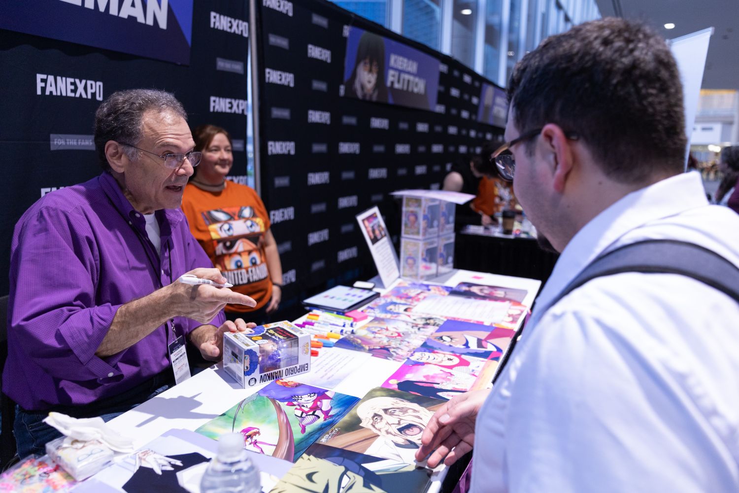 Voice actor Randy Pearlman, wearing a purple shirt, talks animatedly with a fan at his autograph table. Colorful anime prints, markers, and Funko Pop figures are spread across the table, with a FAN EXPO backdrop behind him.