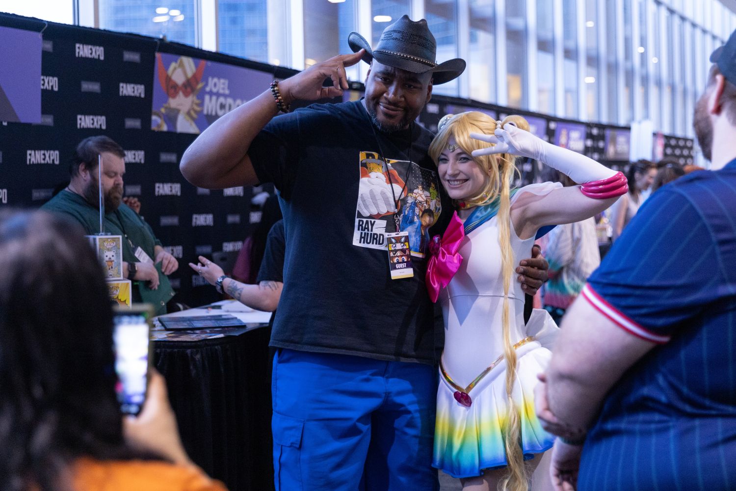 Voice actor Ray Hurd smiles and salutes alongside a fan dressed as Sailor Moon. Both pose with peace signs for a photo, surrounded by autograph tables. A banner for voice actor Joel McDonald is visible in the background.