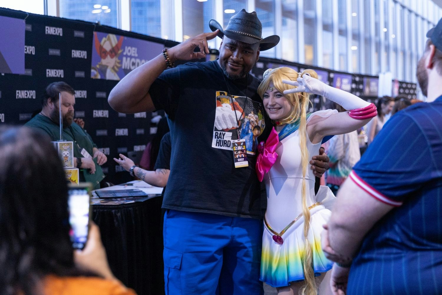 Voice actor Ray Hurd smiles and salutes alongside a fan dressed as Sailor Moon. Both pose with peace signs for a photo, surrounded by autograph tables. A banner for voice actor Joel McDonald is visible in the background.