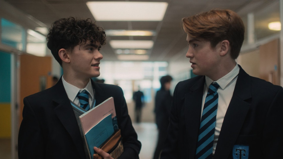 Two of the main characters from Heartstopper, both standing in a hallway inside their school. On the left is Charlie Spring (Joe Locke) and on the right is Nick Nelson (Kit Connor).