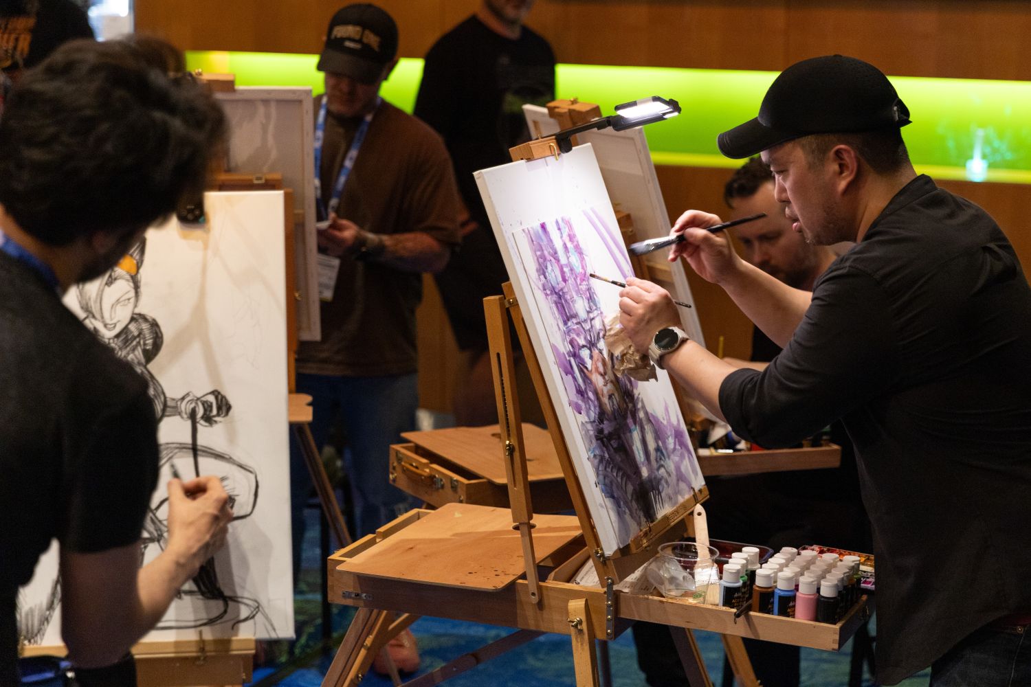 A comic artist in a black shirt and cap paints a vivid purple-toned piece on a canvas while another artist sketches beside him. Their easels are set up in a circle under warm lights as attendees observe in the background.