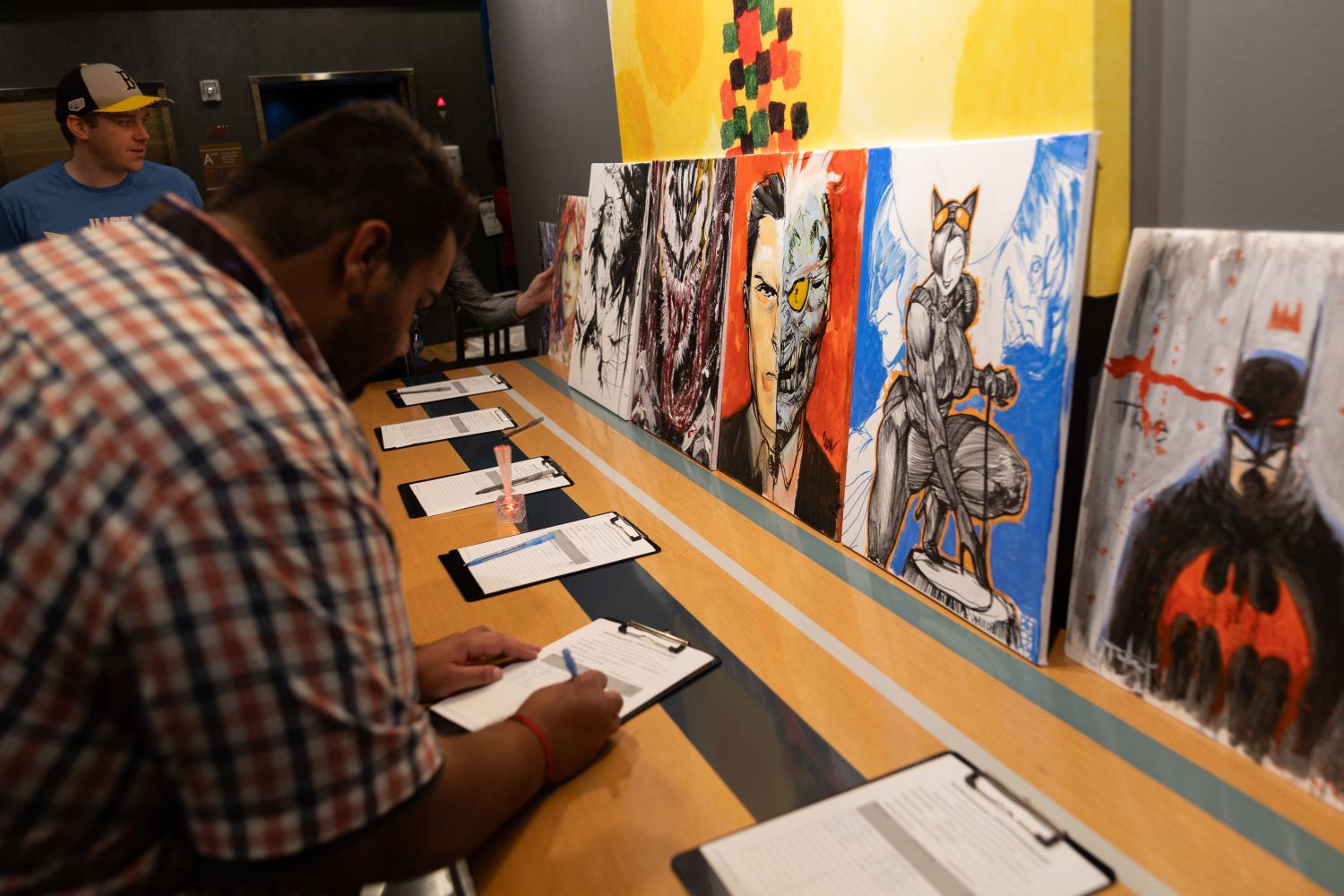 A guest fills out a bid sheet at a silent auction table lined with completed comic art pieces. The displayed artwork includes characters like Venom, Two-Face, Catwoman, and Batman, each painted in bold, stylized forms.