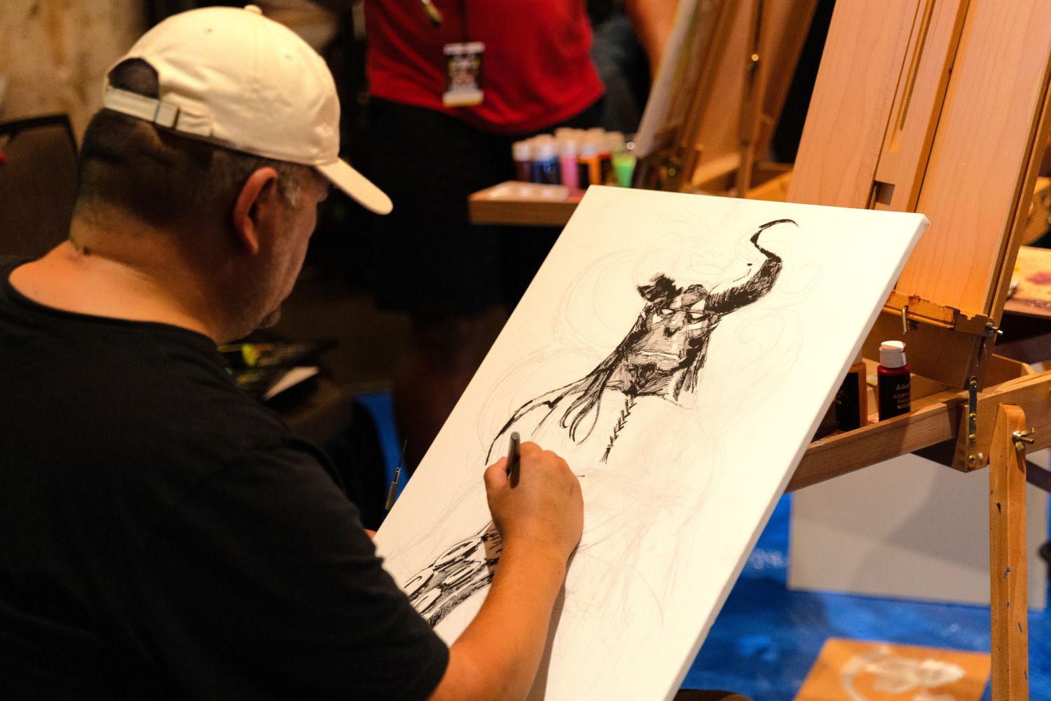 An artist in a white ballcap sketches a dark fantasy-style figure with horns and tattered clothing on a blank canvas. Bottles of acrylic paint are arranged neatly on the tray of the easel beside him.