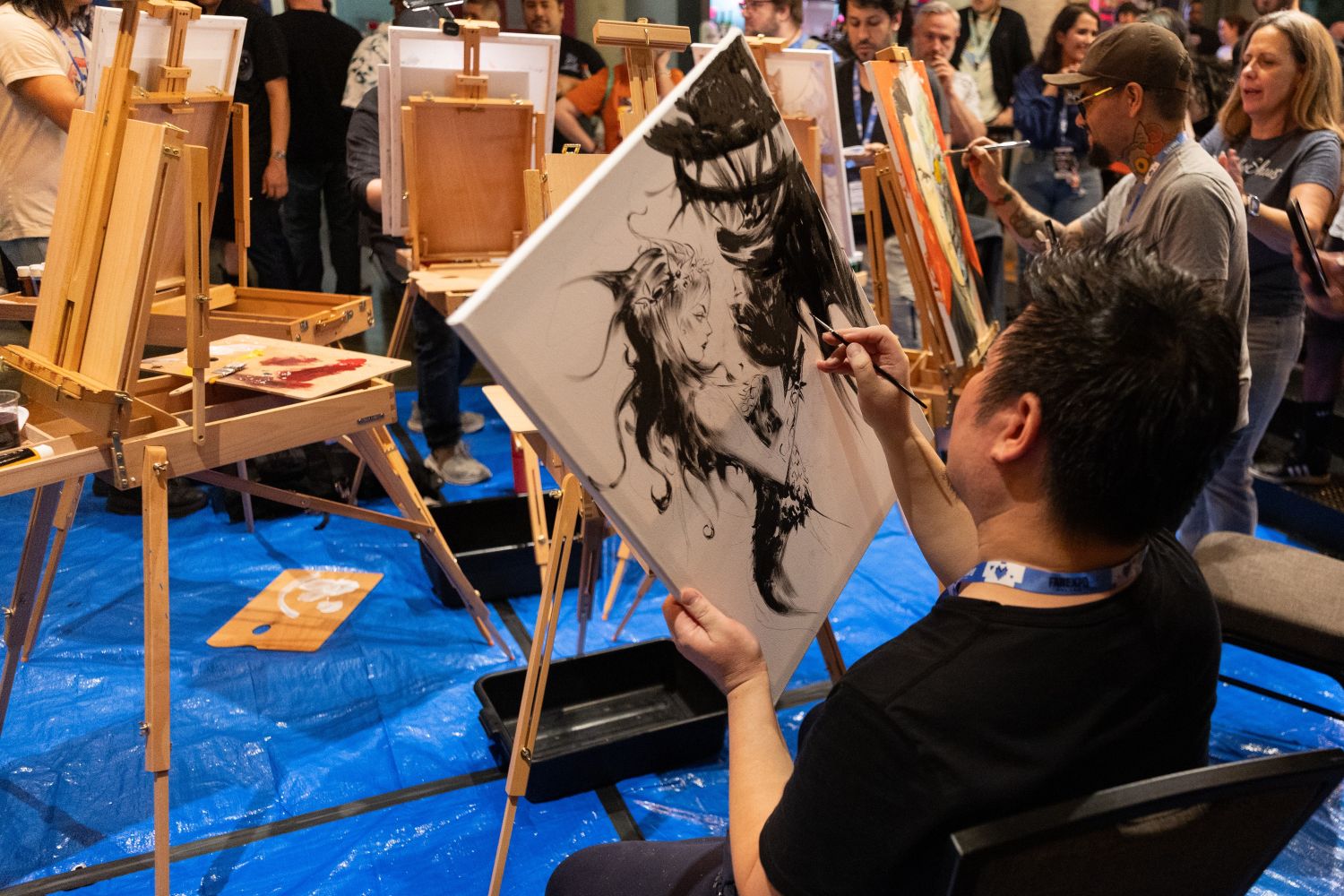 An artist works intently on a dramatic black-and-white illustration of a fantasy female character facing Venom. Other artists are painting at easels nearby, with onlookers watching from behind.