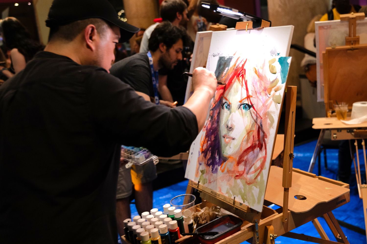A comic artist adds finishing touches to a colorful watercolor portrait of a woman with striking red hair and piercing blue eyes. Paint tubes and brushes are scattered across the supply tray, with other artists working in the background.