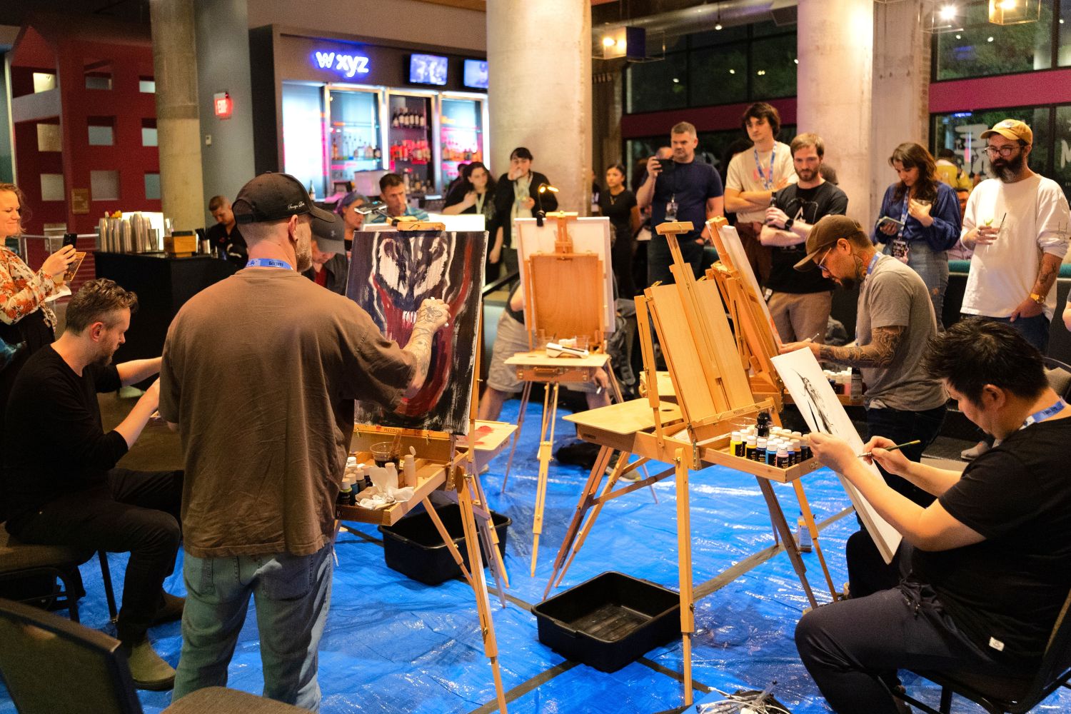 A group of comic artists sit at easels in a lively lounge space, painting live in front of a crowd. One artist works on a menacing close-up of Venom, while others sketch or ink beside him. Guests gather around with drinks, watching and snapping photos.