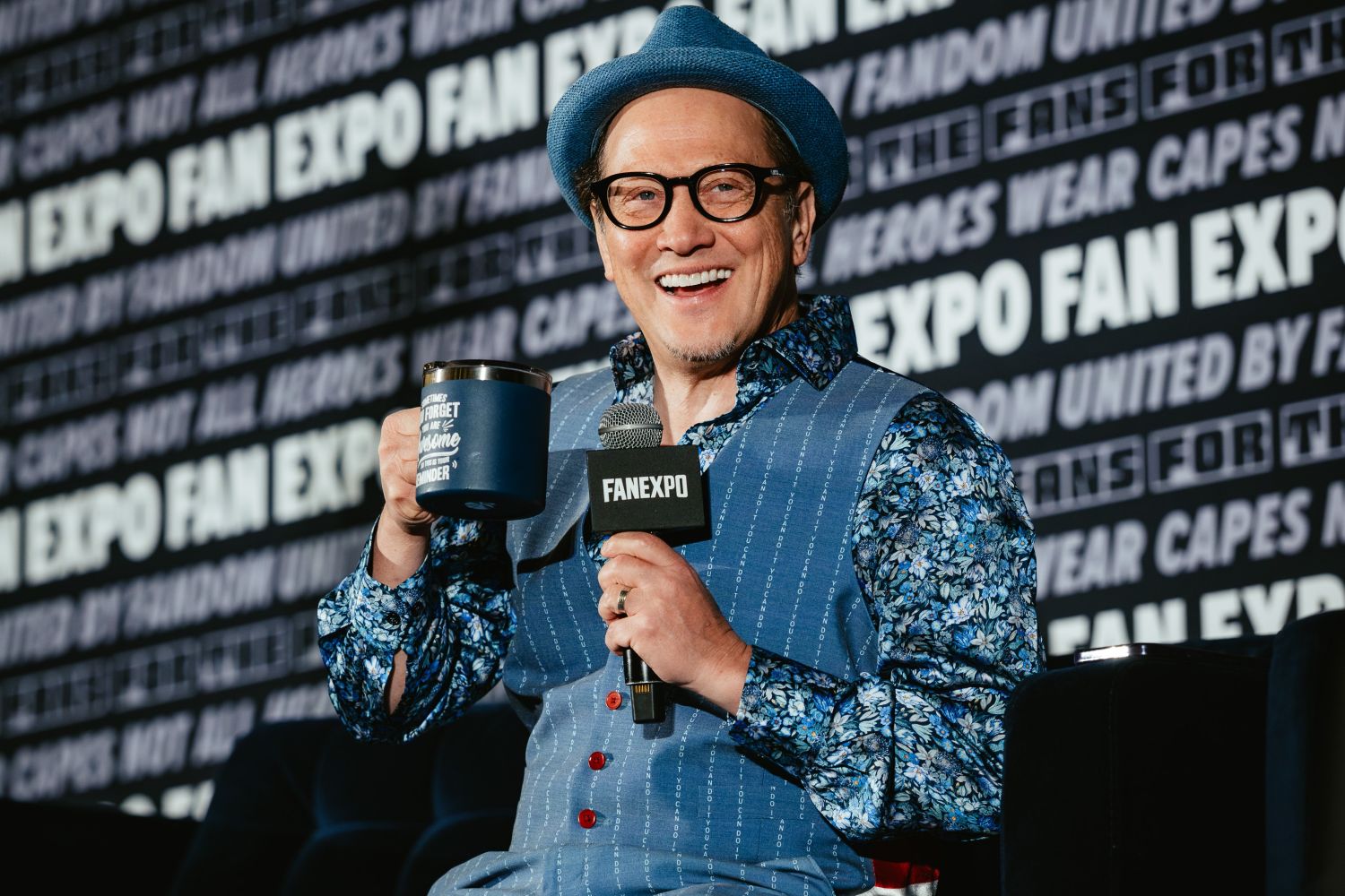 Rob Schneider smiles widely on stage, holding a FAN EXPO microphone and a blue mug that reads “Don’t Forget to Be Awesome.” He wears a blue floral shirt, matching vest, and hat.
