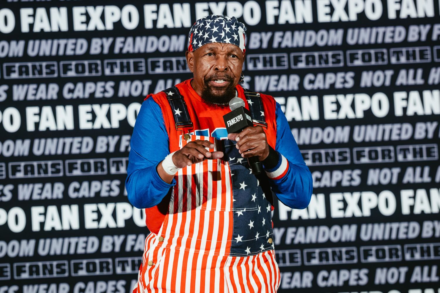 Mr. T stands mid-sentence holding a FAN EXPO mic, dressed in his signature red, white, and blue American flag overalls and headband.