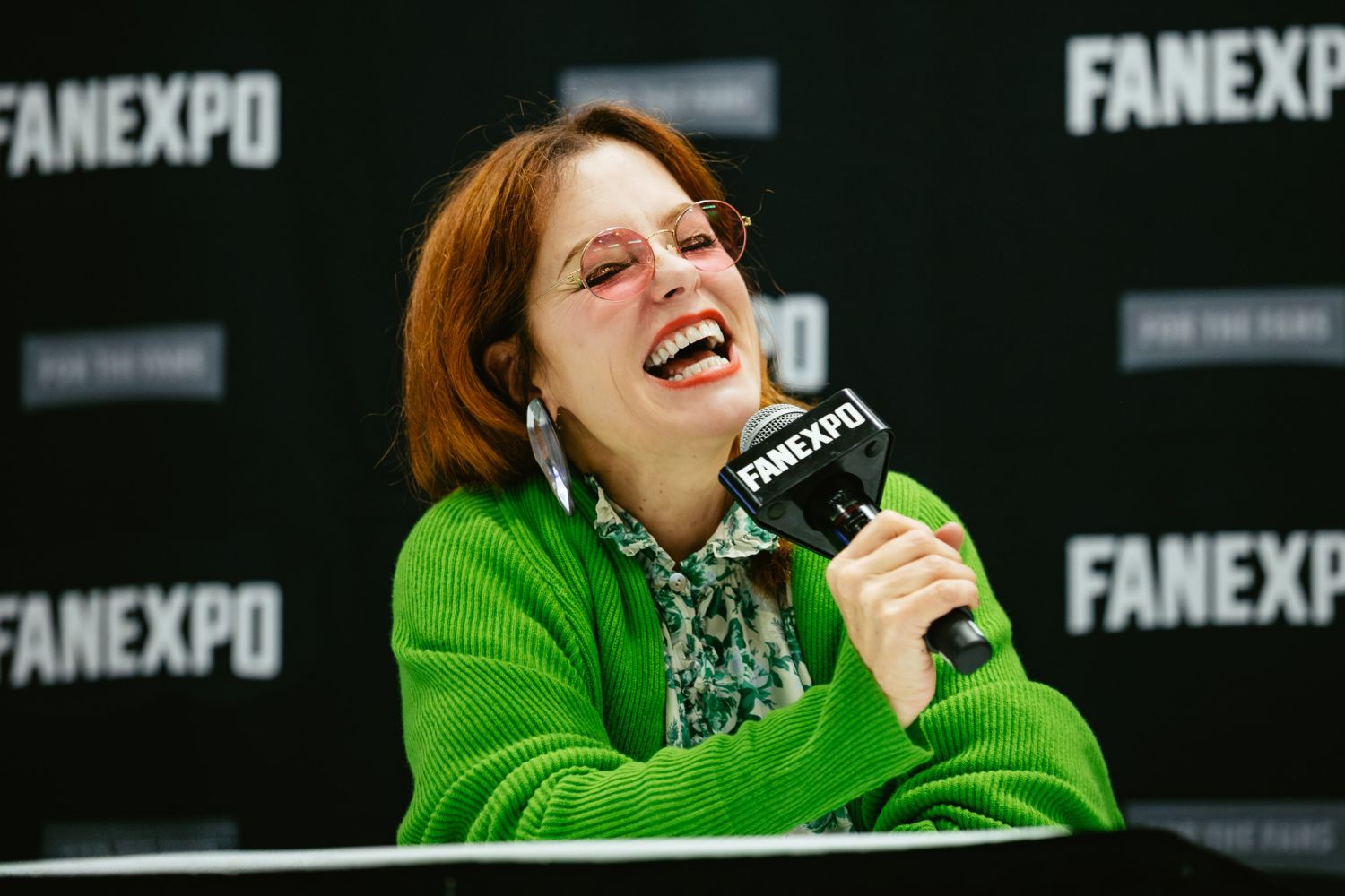 Parker Posey laughs brightly while speaking at her panel, wearing a green cardigan, floral shirt, pink-tinted glasses, and bold earrings.