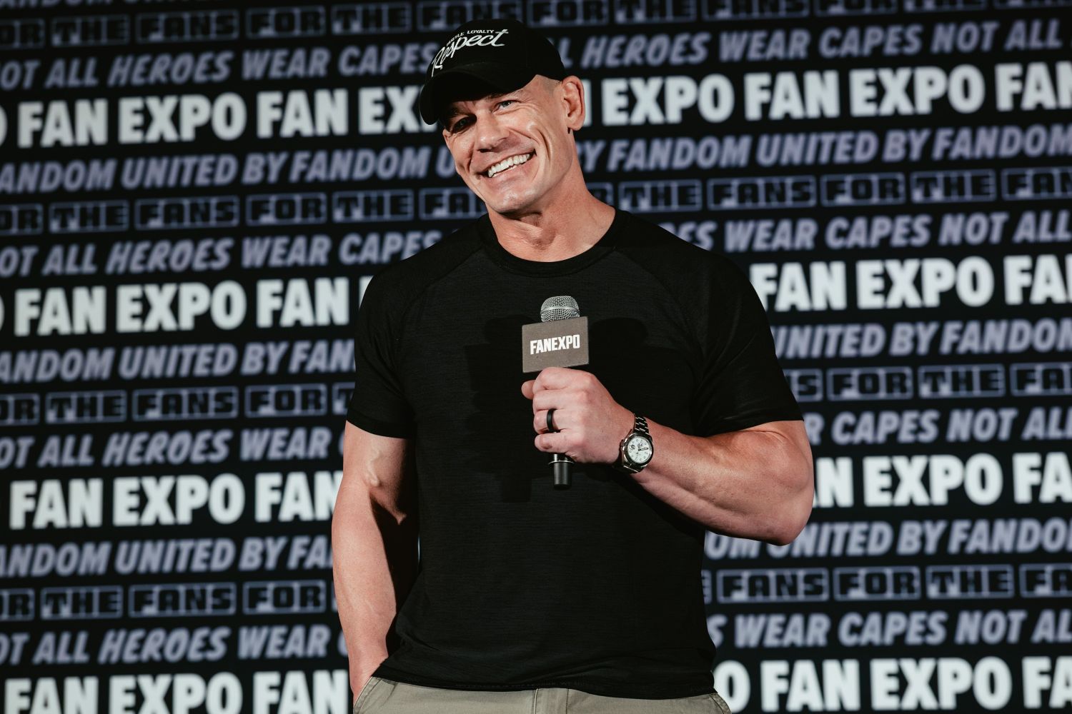 John Cena stands on stage in a black t-shirt and cap, smiling warmly with a FAN EXPO mic in hand, against a branded FAN EXPO backdrop.
