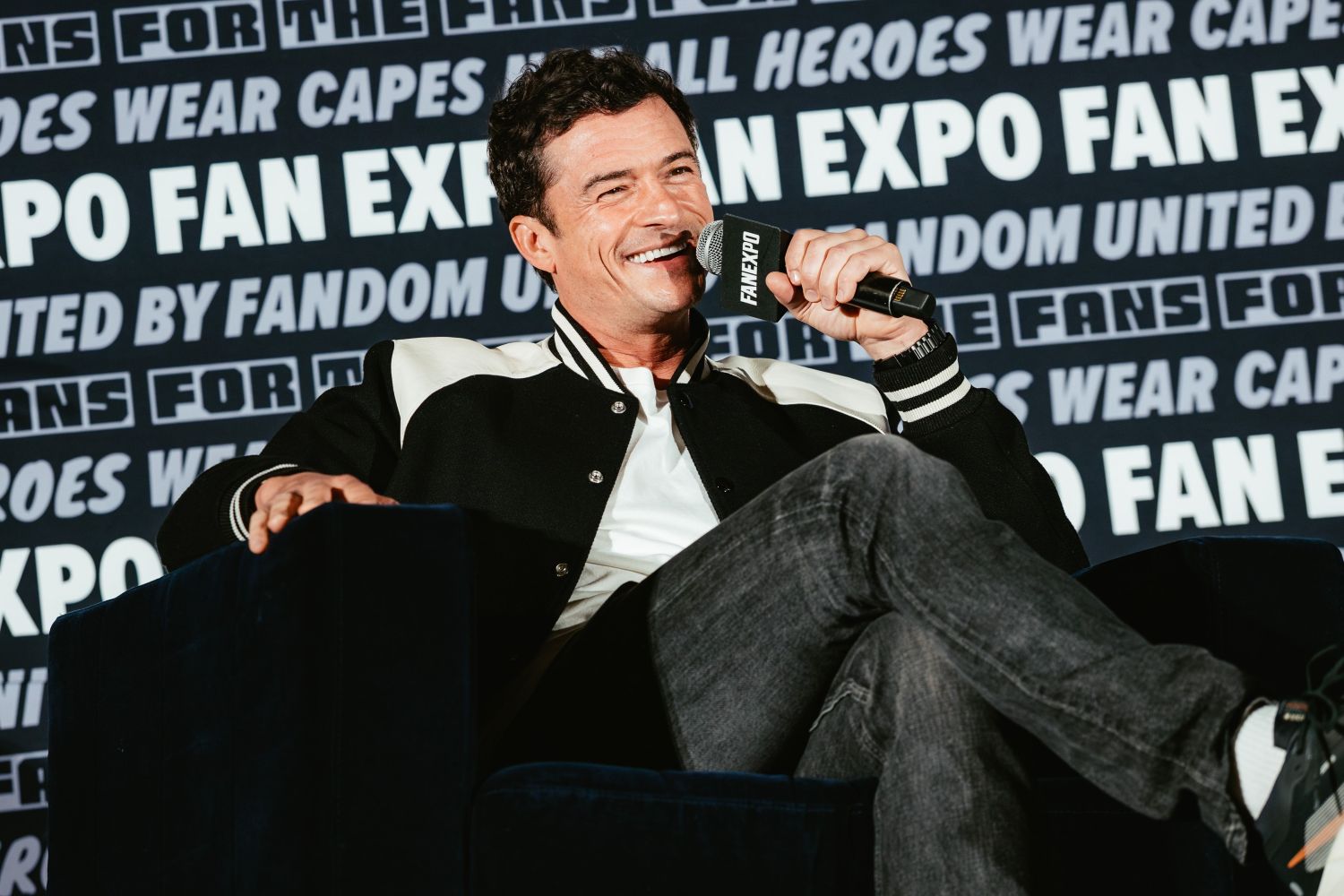 Orlando Bloom lounges in a chair, smiling and speaking into a FAN EXPO mic. He wears a black and white varsity jacket and dark jeans.