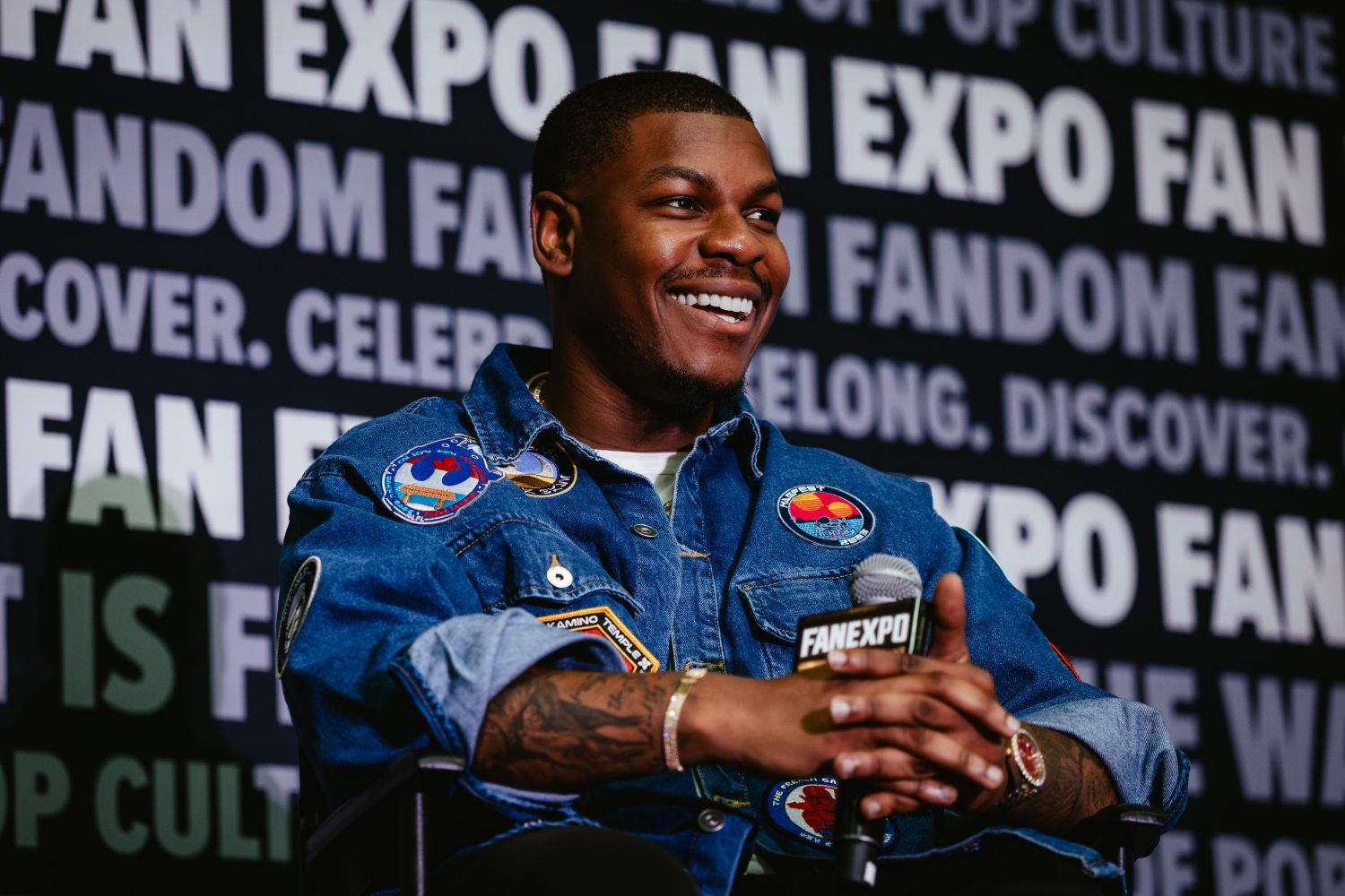 John Boyega smiles as he answers a fan question during his panel, wearing a denim jacket covered in colorful patches and holding a FAN EXPO mic.