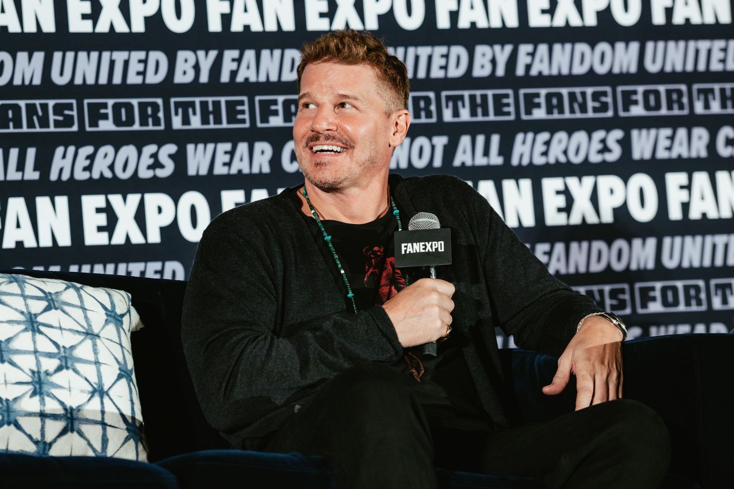 David Boreanaz sits casually with a FAN EXPO mic during his panel, smiling and wearing a black shirt and cardigan with beaded jewelry.
