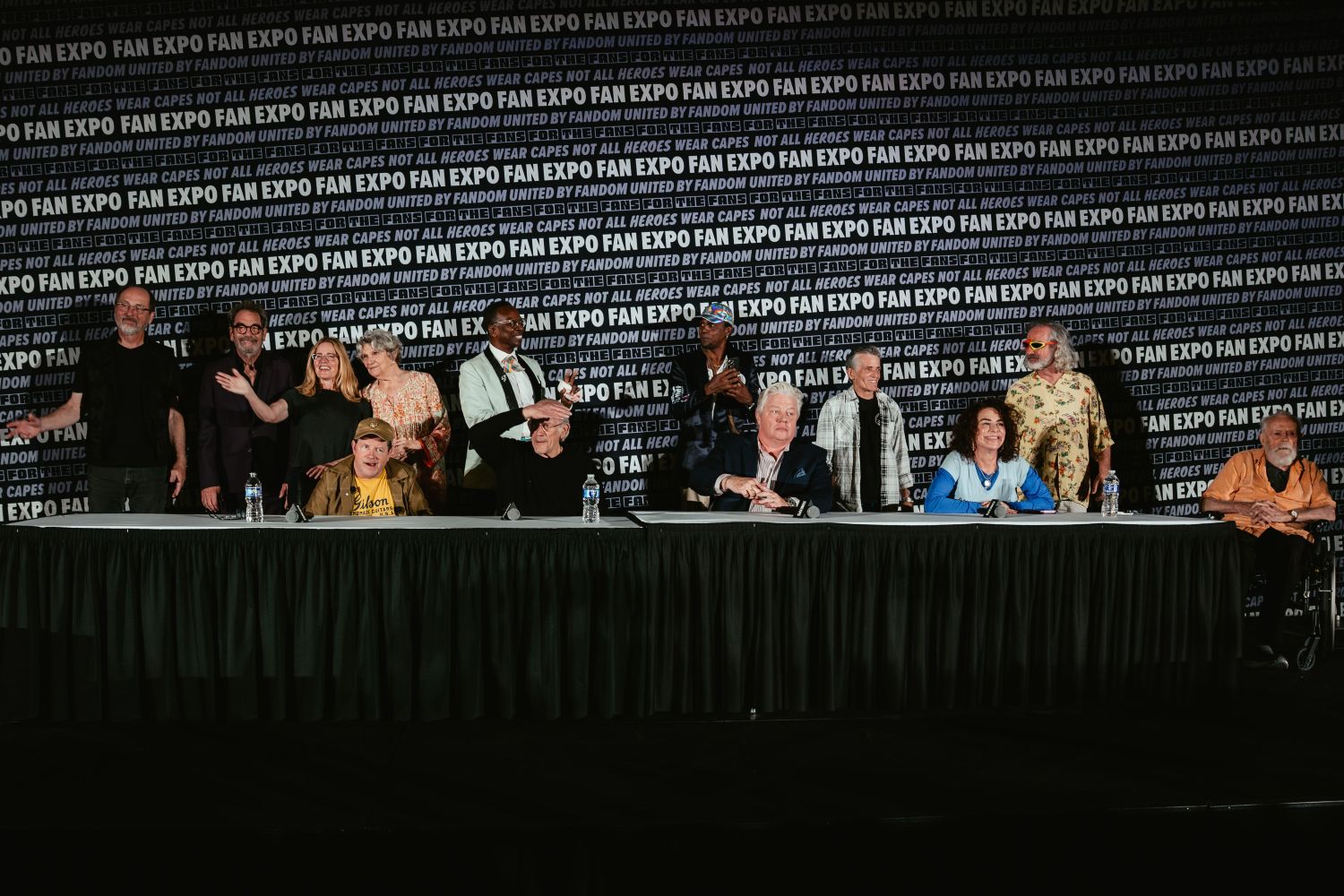A full Back to the Future cast reunion panel features a long table lined with celebrities seated in front of a FAN EXPO backdrop, while others stand behind them, waving and smiling.