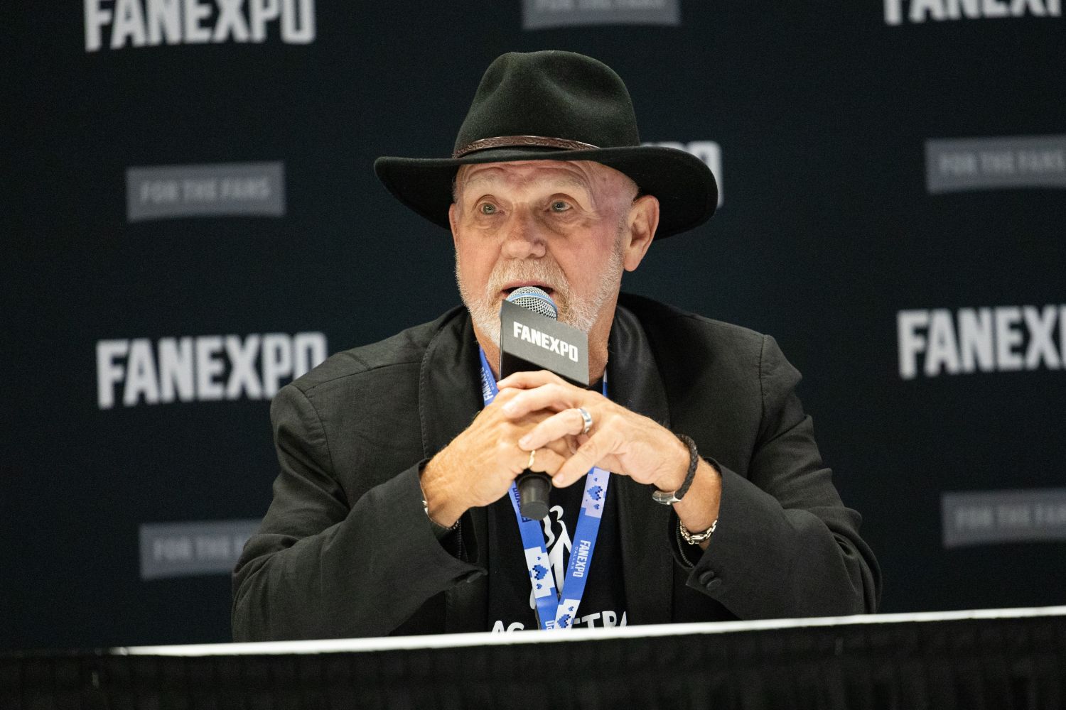 Steve Downes, in a black blazer and cowboy hat, sits at a table during a FAN EXPO panel, speaking into a microphone with a serious expression.