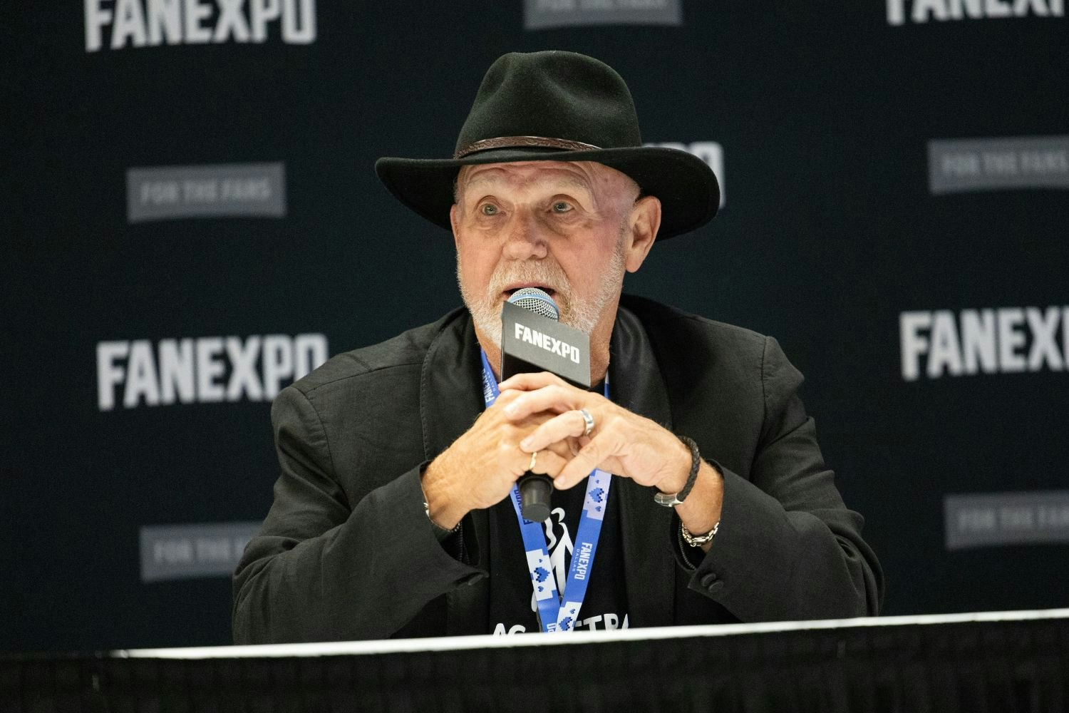 Steve Downes, in a black blazer and cowboy hat, sits at a table during a FAN EXPO panel, speaking into a microphone with a serious expression.