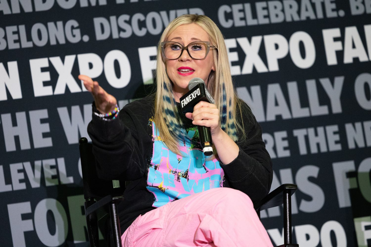 Ashley Eckstein, with long blond hair and pink-accented glasses, gestures animatedly while speaking into a FAN EXPO mic. She wears a black hoodie and pink pants, with colorful hair and accessories.