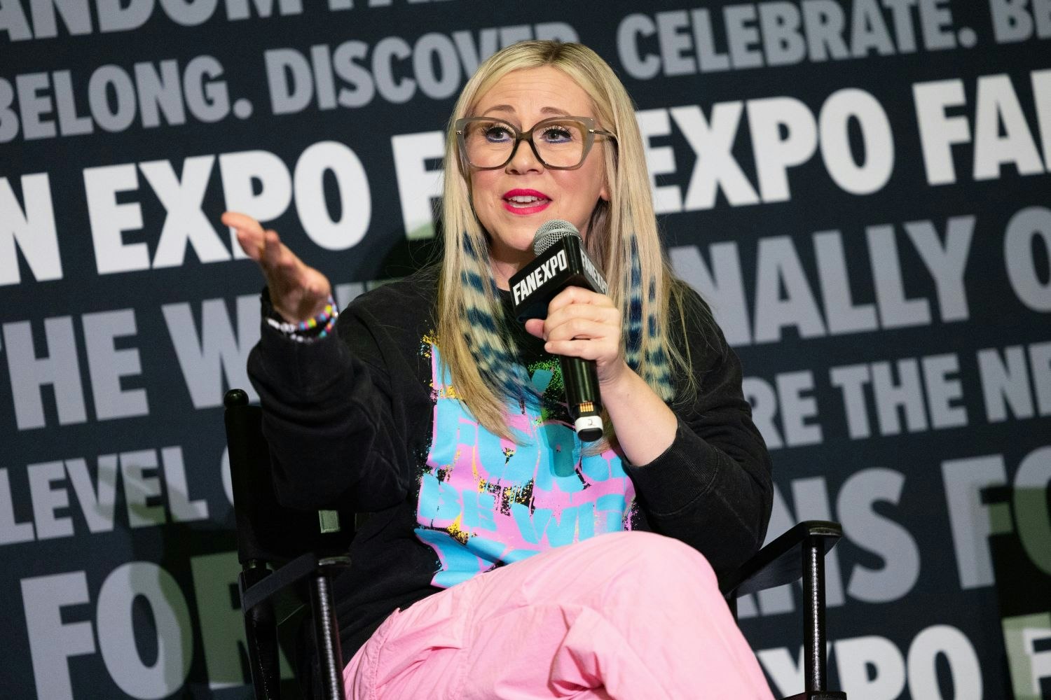 Ashley Eckstein, with long blond hair and pink-accented glasses, gestures animatedly while speaking into a FAN EXPO mic. She wears a black hoodie and pink pants, with colorful hair and accessories.